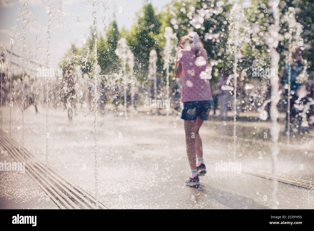 Peu cute girl walking in open street fontaine à jour ensoleillé chaud. Vue arrière. Banque D'Images