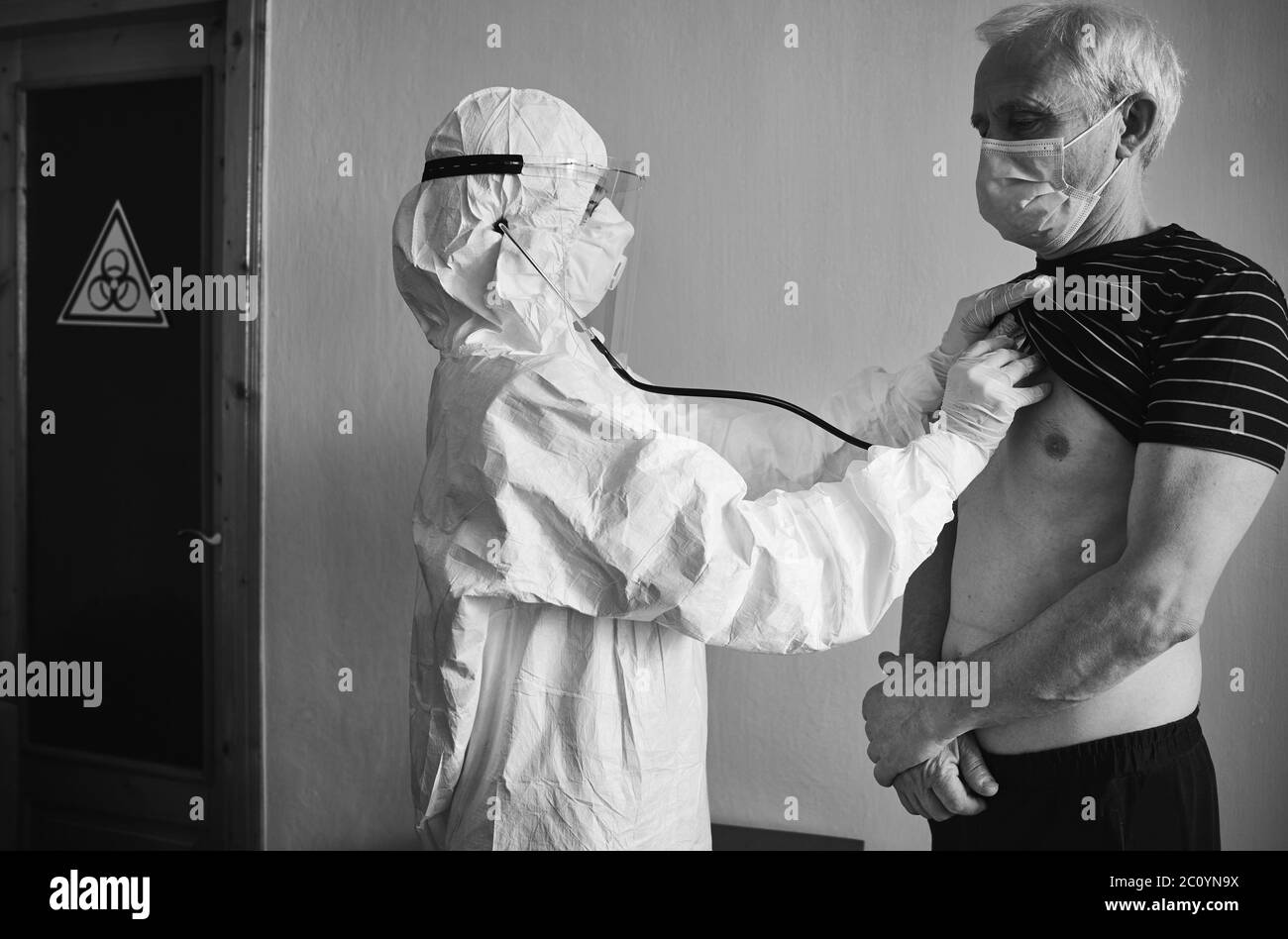 Médecin en costume de protection personnelle examinant le patient âgé de sexe masculin à la maison à l'aide d'un phonendoscope, en vérifiant la poitrine, debout au mur, panneau de danger biologique sur la porte, noir et blanc, concept COVID-19 Banque D'Images
