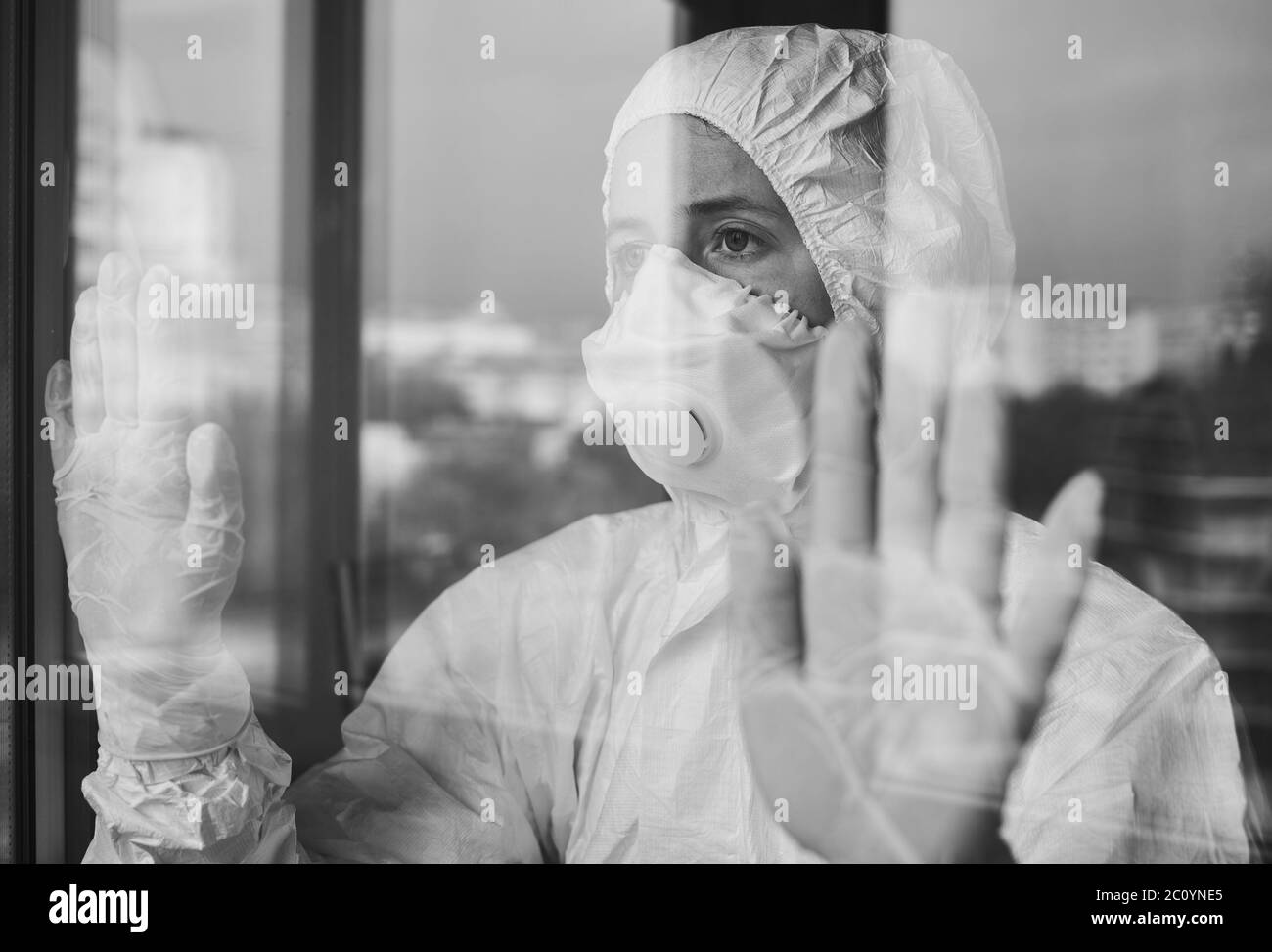 Portrait rapproché d'une femme malheureuse à l'auto-isolation, portant un vêtement d'EPI, un respirateur, des gants, tout en se tenant à la maison et regardant par la fenêtre. Concept de coronavirus Banque D'Images