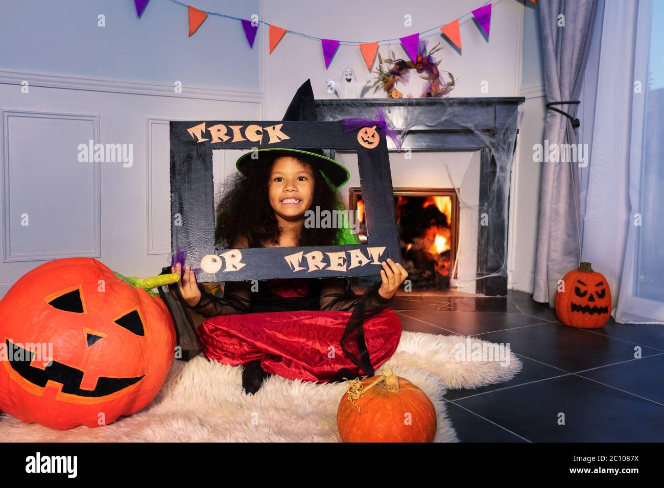 Fille avec un cadre de trick ou de traite dans la robe d'Halloween s'asseoir à la maison près de la cheminée et de grandes citrouilles d'orange Banque D'Images