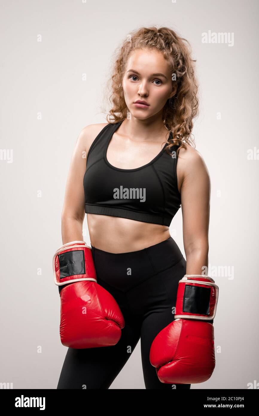 Blonde FIT fille en survêtement noir et gants de boxe rouges debout devant l'appareil photo en isolation prêt pour l'entraînement dans un club de sport ou une salle de gym Banque D'Images