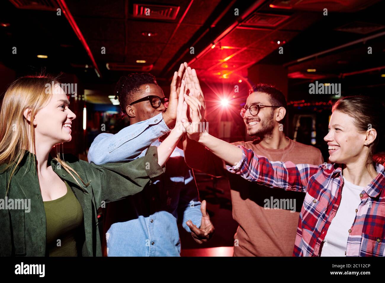 Deux jeunes couples interculturels heureux en vêtements décontractés se donnant les uns les autres haut cinq après partie de bowling dans le centre de divertissement Banque D'Images