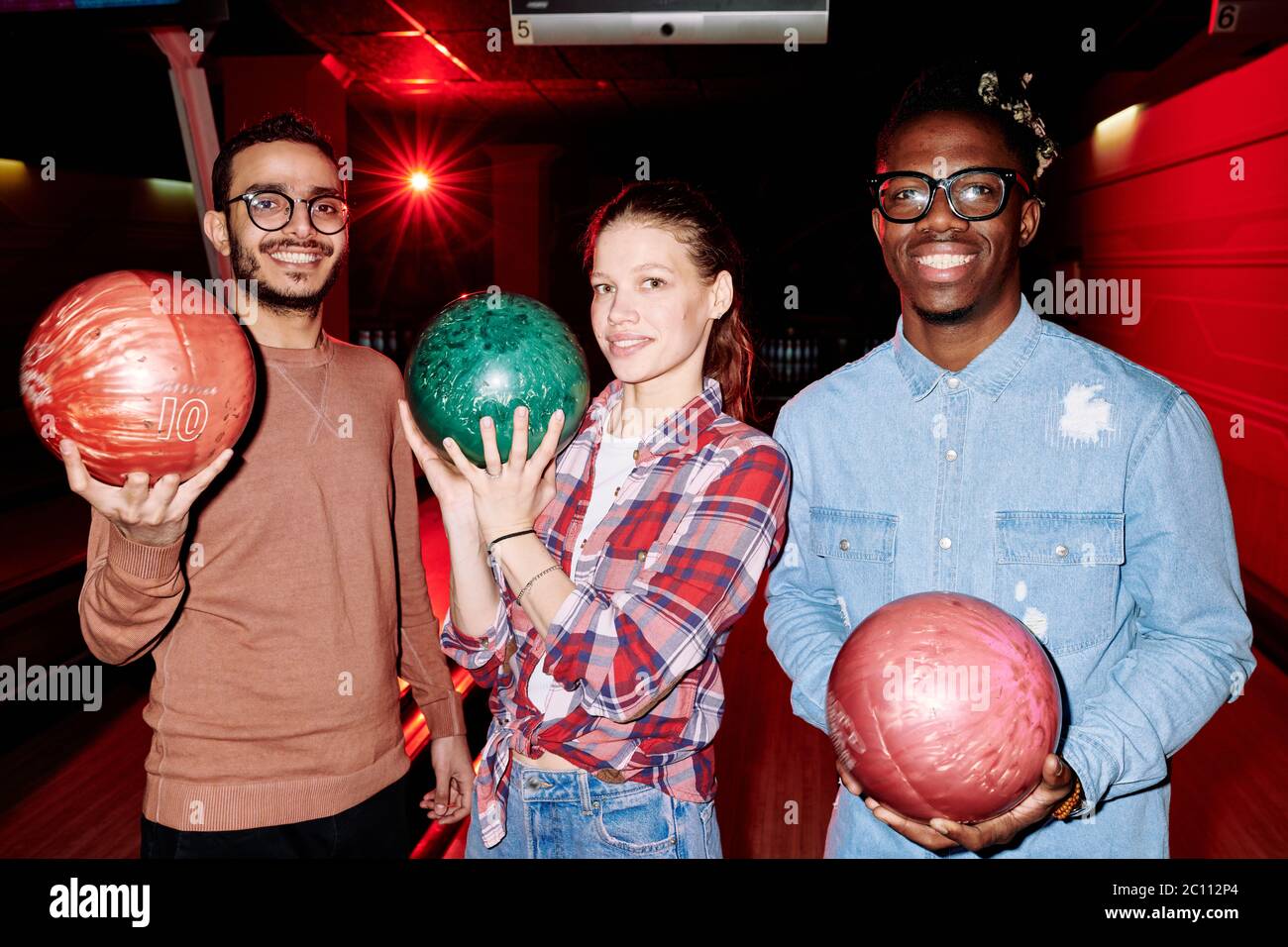 Jolie jeune femme et deux types interculturels avec des sourires crasseux tenant des boules de bowling tout en jouant ensemble dans un club de divertissement Banque D'Images