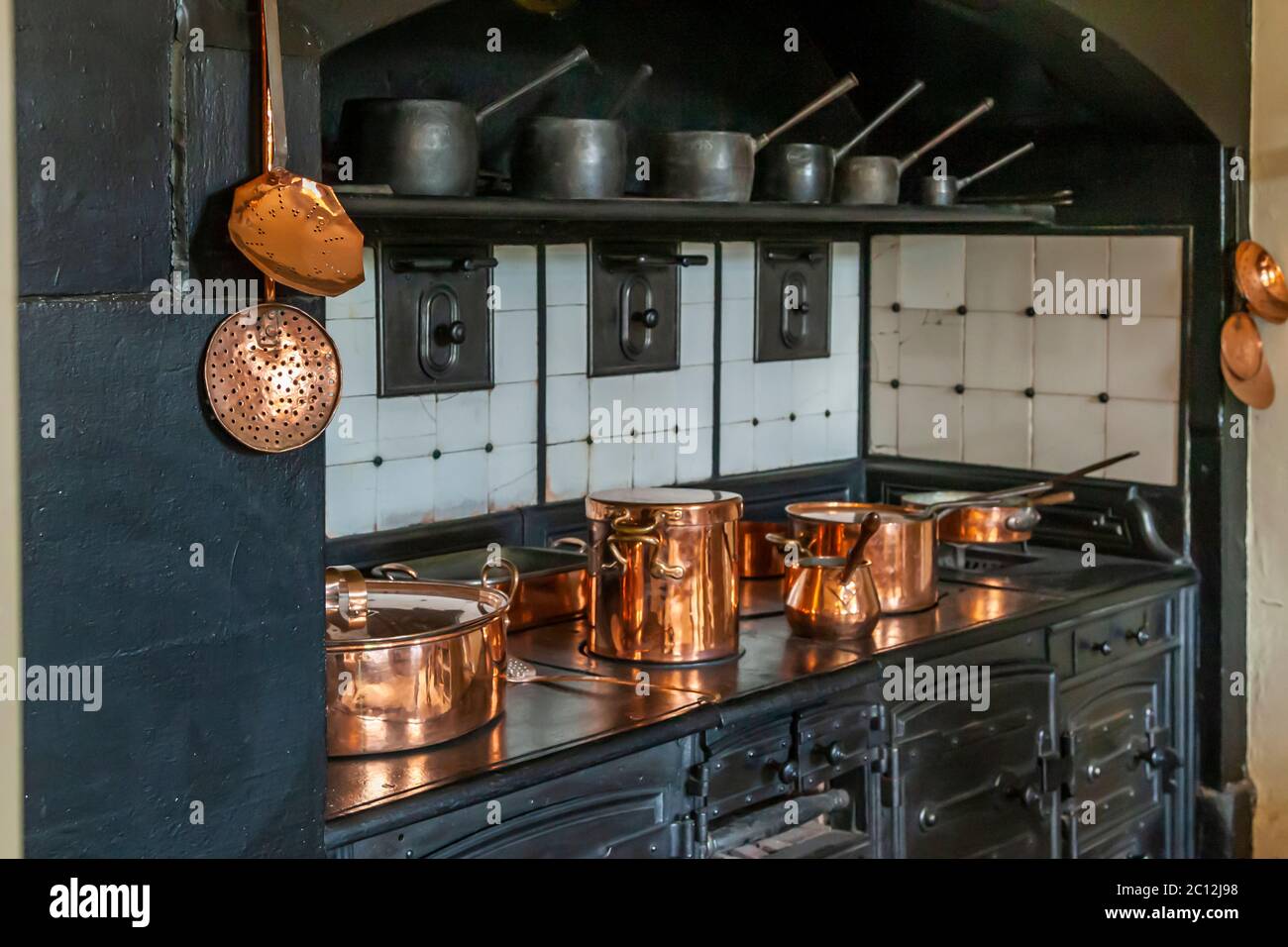 Plusieurs casseroles en cuivre de tailles différentes brillent à la lumière sur la cuisinière noire dans la cuisine d'Audley End House près de Saffron Walden, Essex, Angleterre. Une petite cruche en cuivre avec une poignée en bois se trouve parmi les plus grands pots à couvercle. Thomas Howard, 1er comte de Suffolk, a construit Audley End House entre 1605 et 1614 dans le style jacobéen, autrefois l'une des plus grandes maisons de campagne d'Angleterre. English Heritage gère le domaine depuis 1984. Banque D'Images
