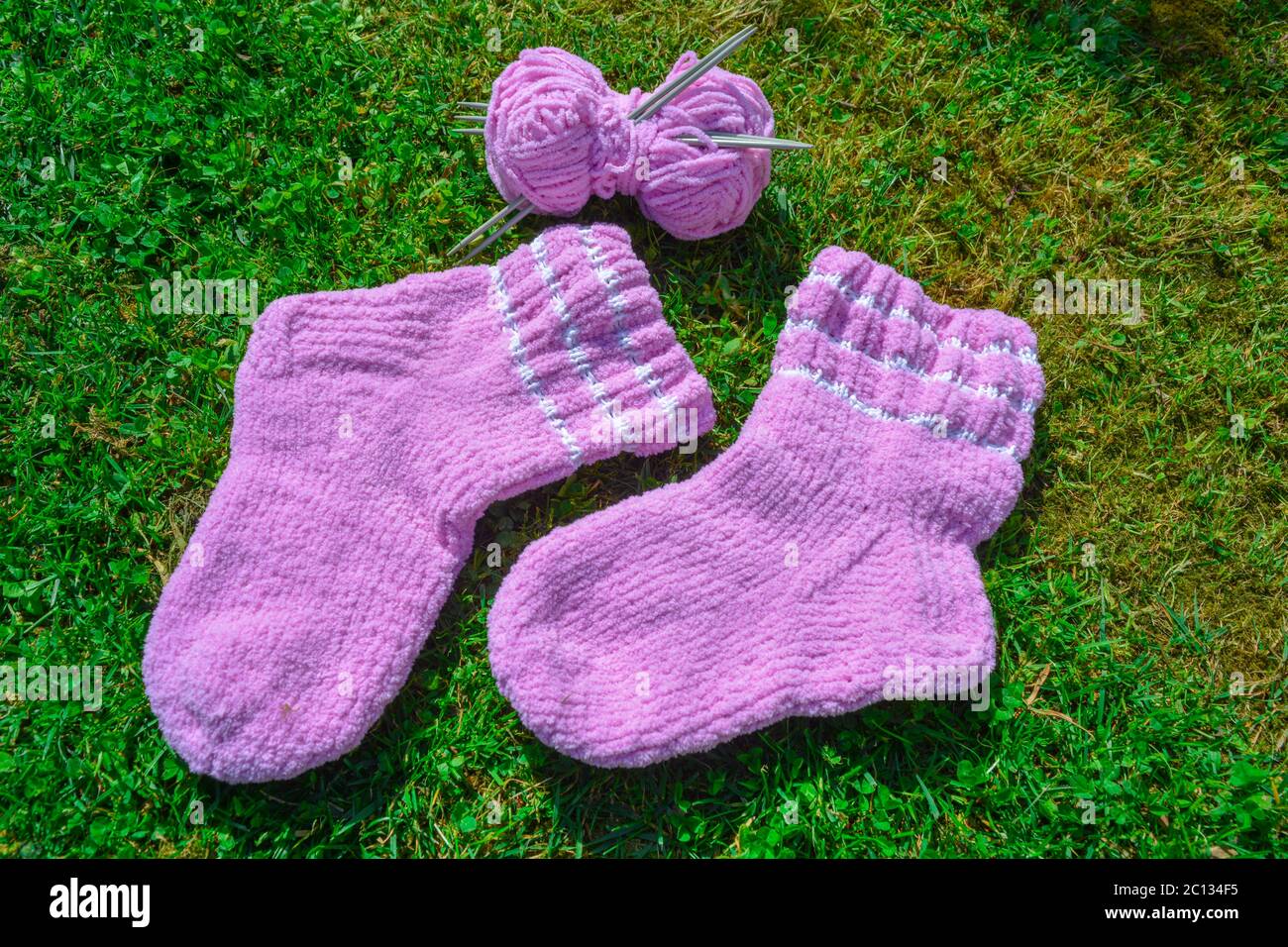 Une paire de chaussettes en laine rose et une balle de fil avec un ensemble d'aiguilles sur fond d'herbe verte. Chaussettes synthétiques faites à la main avec rayures blanches, tissu doux Banque D'Images