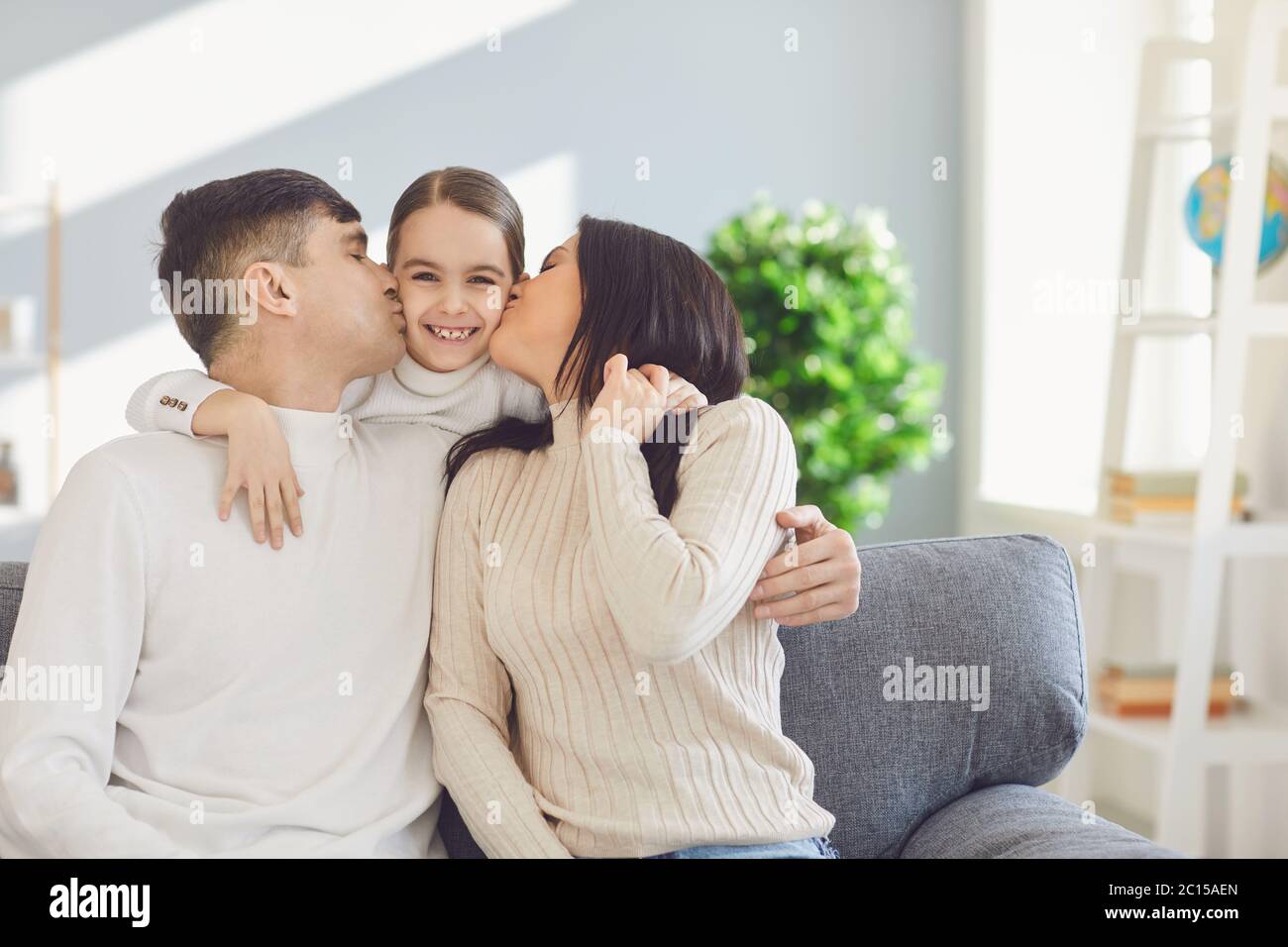 Bonne famille dans la chambre. Mère et père embrassent une fille assise sur un canapé dans une pièce lumineuse et ensoleillée à la maison Banque D'Images