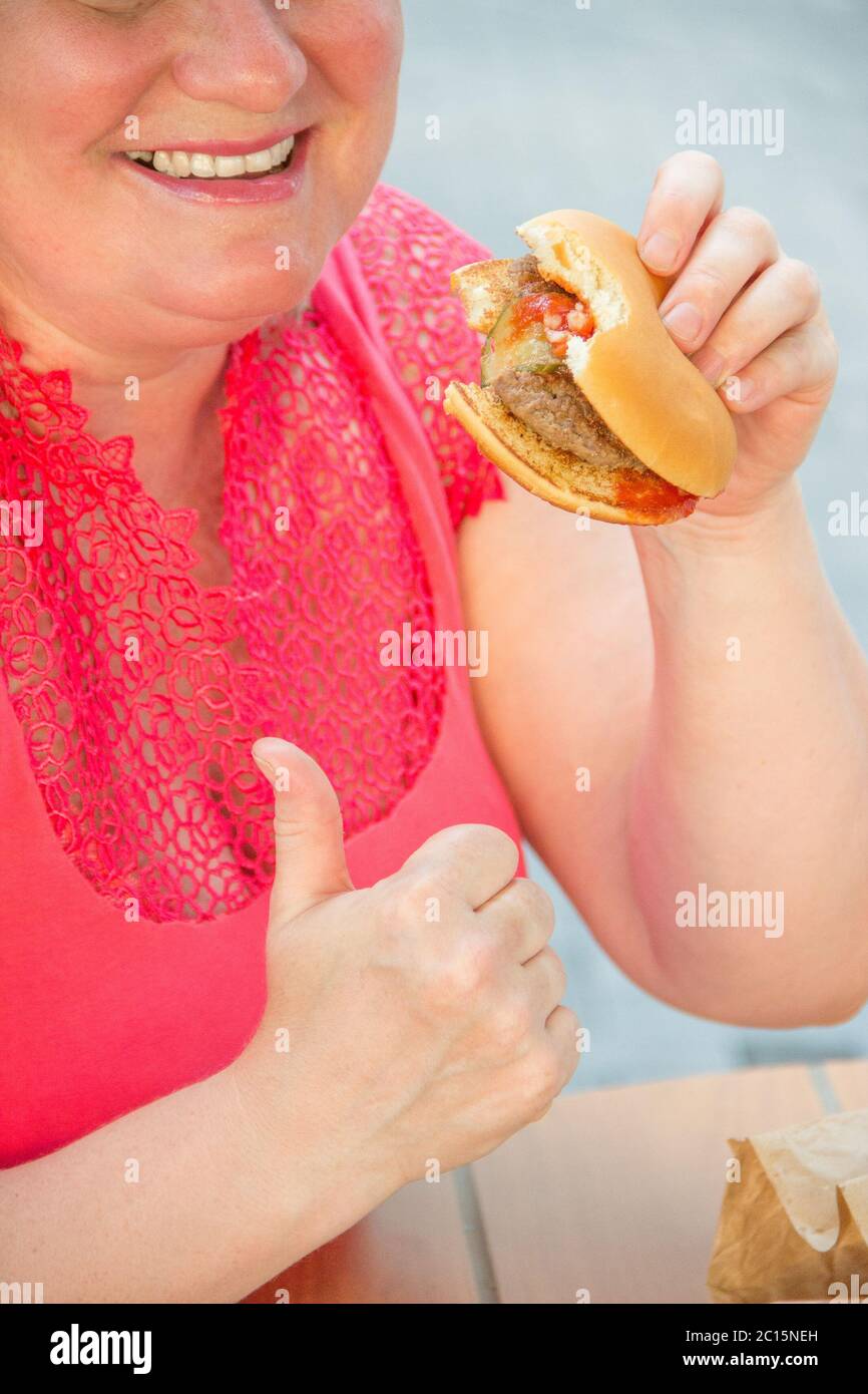 Femme épaisse en vêtements rouges mangeant un hamburger fast food et des frites dans un café à une table dans une rue d'été Banque D'Images