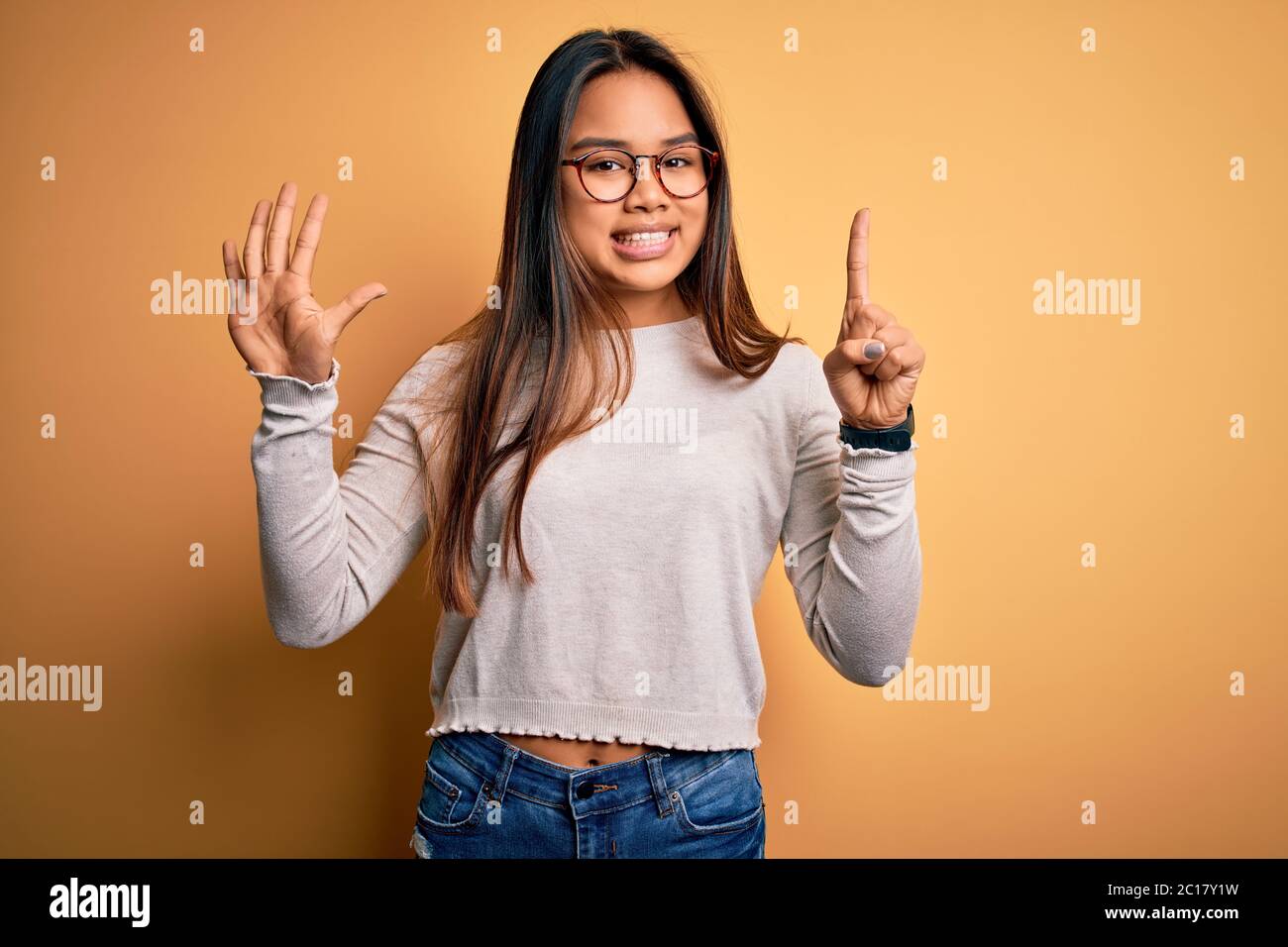 Jeune fille asiatique belle portant un chandail décontracté et des lunettes sur fond jaune montrant et pointant vers le haut avec les doigts numéro six tout en souriant con Banque D'Images