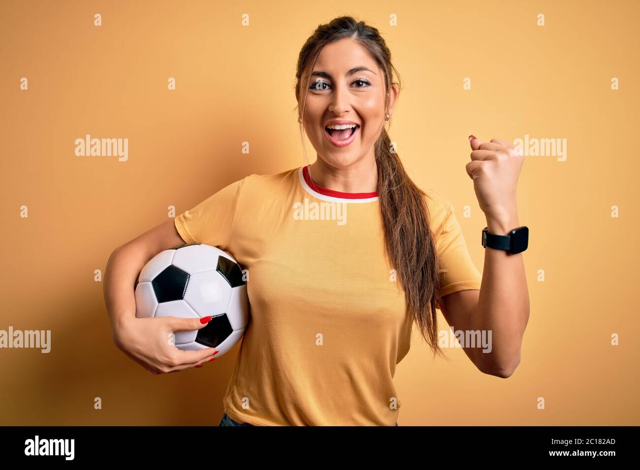 Belle brunette joueur femme jouant au football en utilisant le ballon de football sur fond jaune criant fier et célébrant la victoire et le succès très exc Banque D'Images