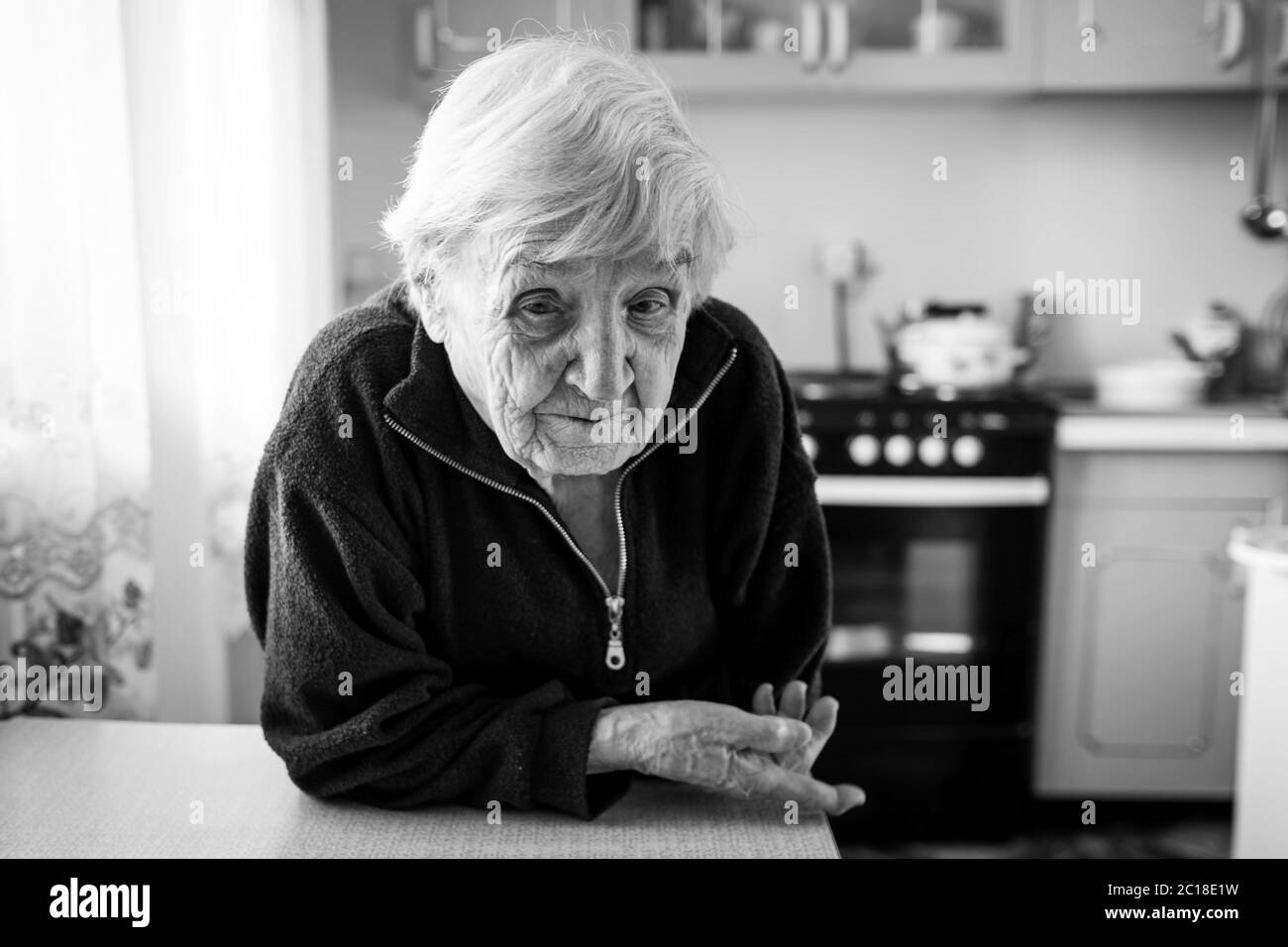 Une vieille femme dans la cuisine près de la fenêtre de sa maison. Photo en noir et blanc. Banque D'Images