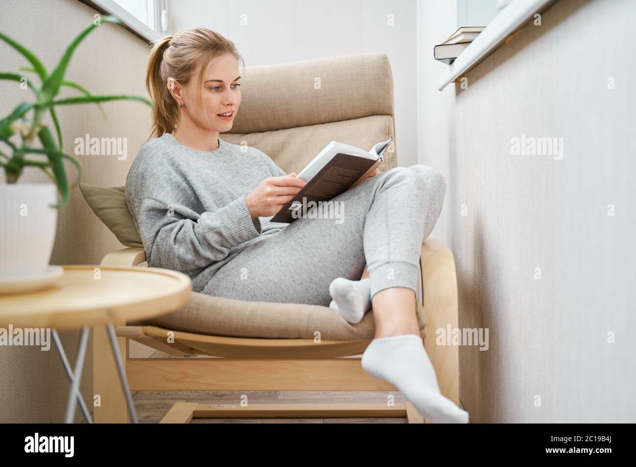 Femme blonde lisant le livre tout en étant assise sur un fauteuil beige appartement Banque D'Images