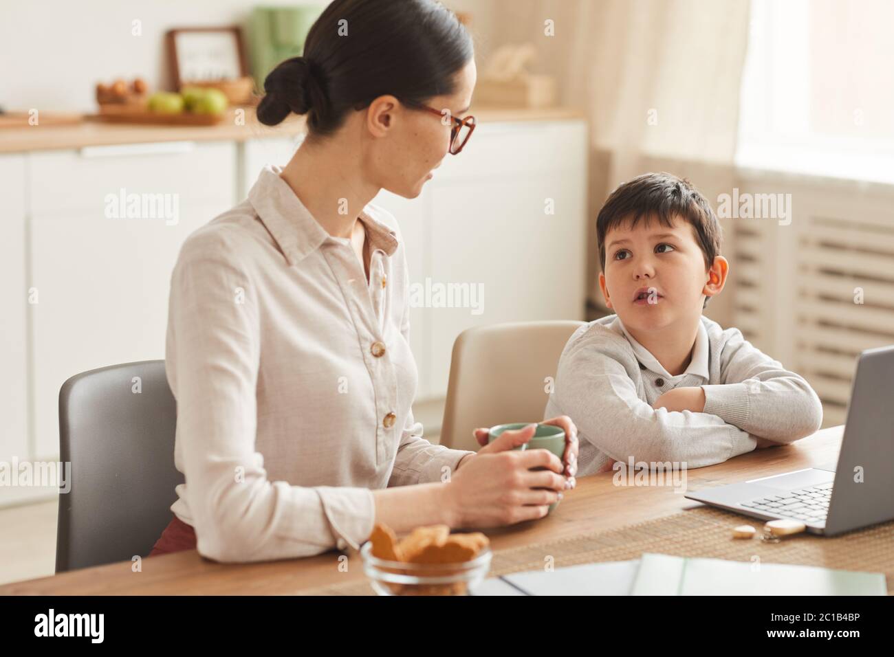 Portrait aux tons chaleureux d'une jeune mère qui parle à son fils tout en faisant ses devoirs ensemble assis à une table dans un intérieur confortable de cuisine, espace pour les copies Banque D'Images
