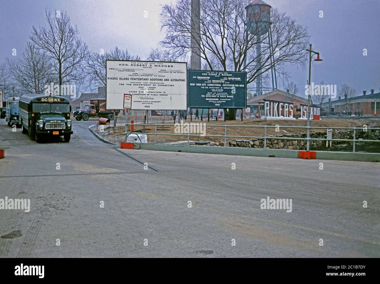 La route menant au complexe pénitentiaire de l'île Rikers par une journée d'hiver, East River, New York, USA 1963. Les grandes lettres 3-D qui orthographient 'Rikers Island' sont à droite, en face de la tour d'eau. Un bus de la prison part (à gauche). Les panneaux indiquent que des travaux de construction importants sont en cours sous la direction du maire de New York Robert F Wagner. Banque D'Images