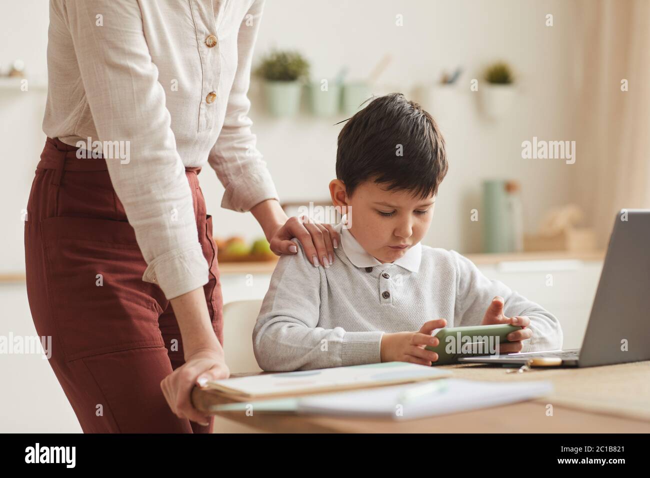 Portrait d'un garçon mignon jouant au jeu mobile tout en étudiant à la maison avec la mère debout près de lui, espace de copie Banque D'Images