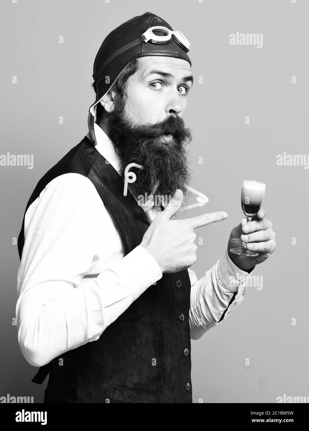 Beau pilote barbu ou aviator homme avec longue barbe et moustache sur drôle de visage holding glass de boissons tourné en vintage cuir gilet avec chapeau, lunettes et sur fond bleu studio Banque D'Images