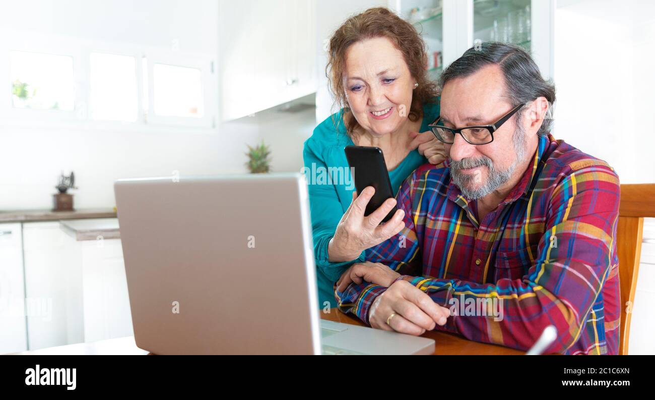 Un couple âgé utilise simultanément son smartphone et son ordinateur portable depuis sa cuisine à la maison. Restez à la maison et nouveau concept de communication. Banque D'Images