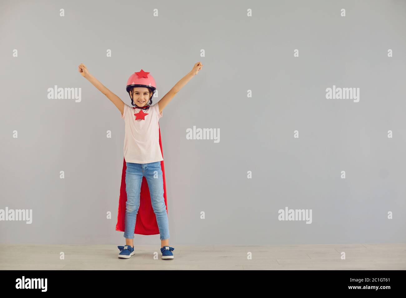 Le petit super-héros de bébé dans un casque sourit ses mains tout en se tenant sur un fond gris. Banque D'Images