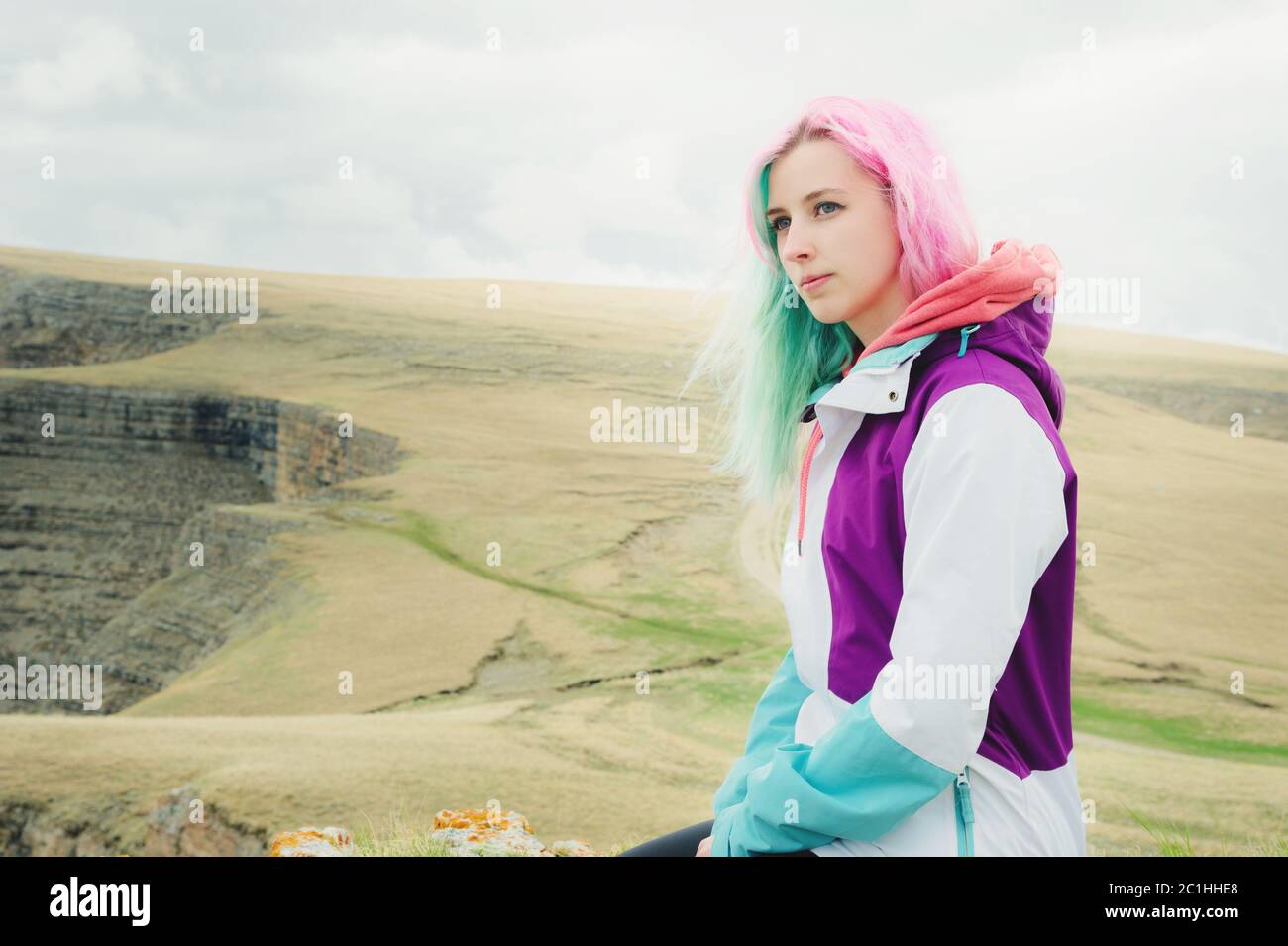 Une jeune fille-voyageur aux cheveux multicolores est assise sur le bord d'une falaise et regarde à l'horizon sur un fond de plateau rocheux Banque D'Images