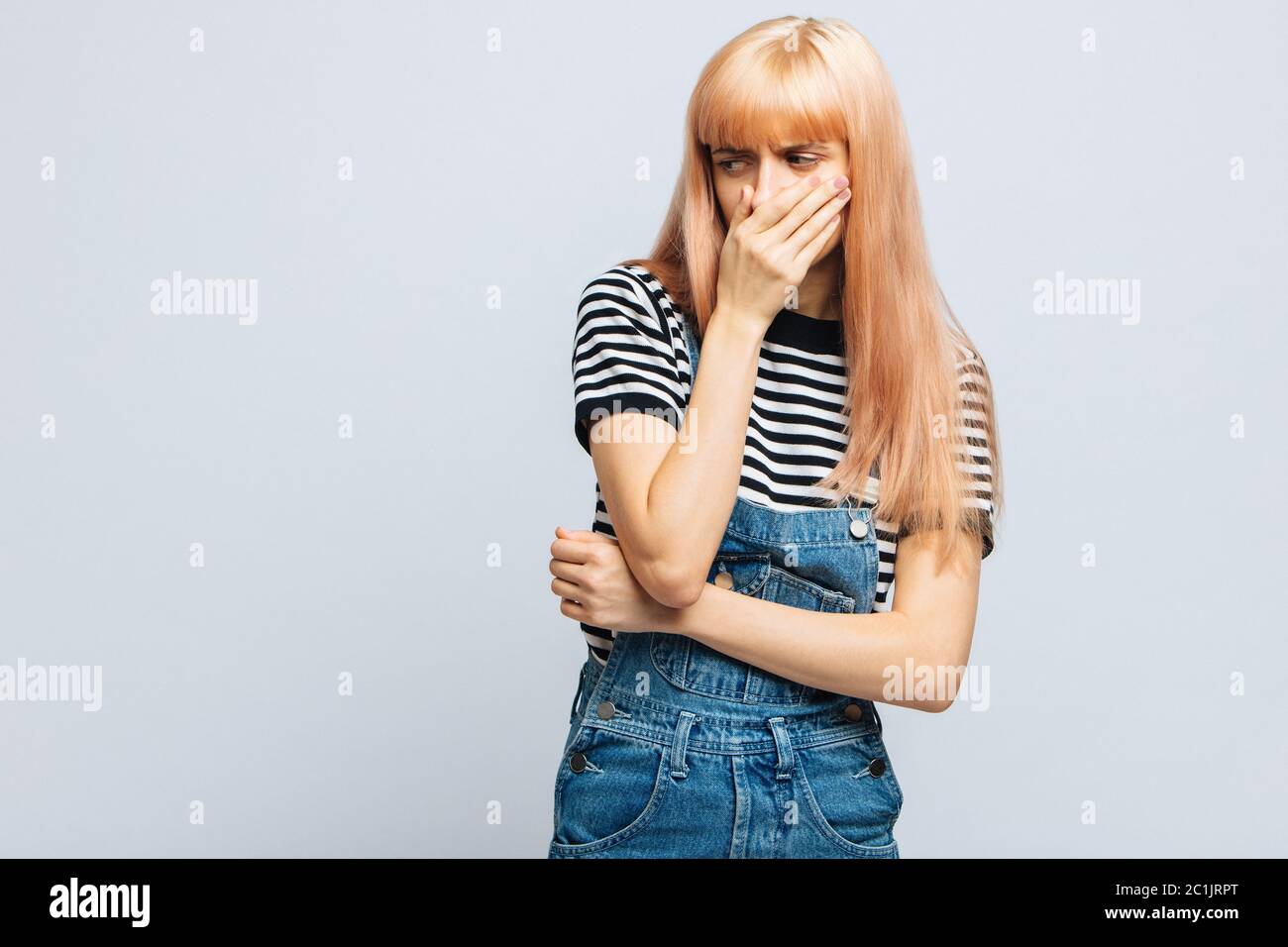 Studio portrait de malsain mignon blond femelle en t-shirt rayé couvre le nez avec la main, regarde à la source de l'allergie ou l'odeur désagréable Banque D'Images