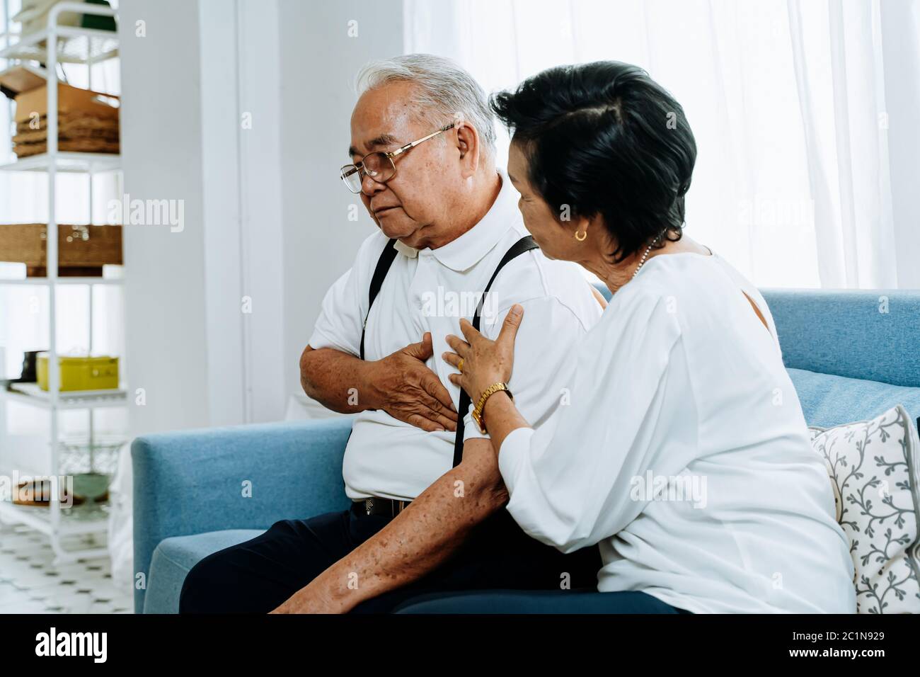 Femme âgée prenant soin de l'homme asiatique âgé souffrant de brûlures d'estomac tout en étant assise sur un canapé dans le salon confortable à la maison. Ancien retraité malade Banque D'Images