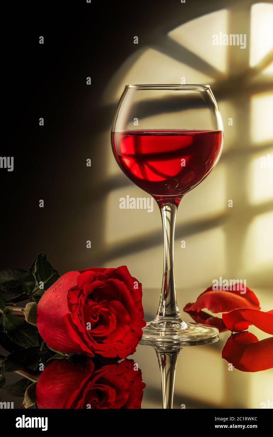 un verre de vin rouge et une rose rouge sur la table avec réflexion, lumière de la fenêtre Banque D'Images