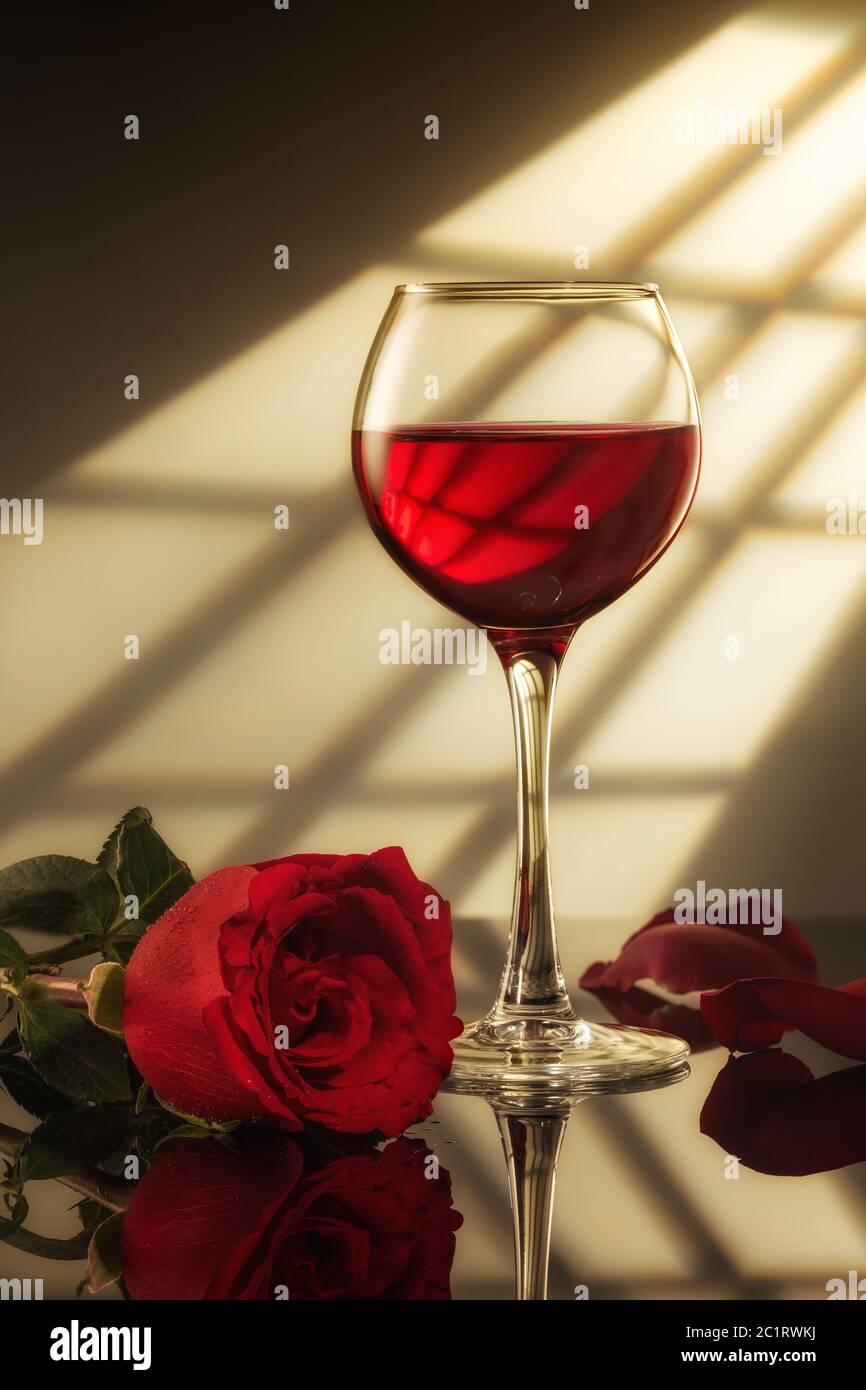 un verre de vin rouge et une rose rouge sur la table avec réflexion, lumière de la fenêtre Banque D'Images