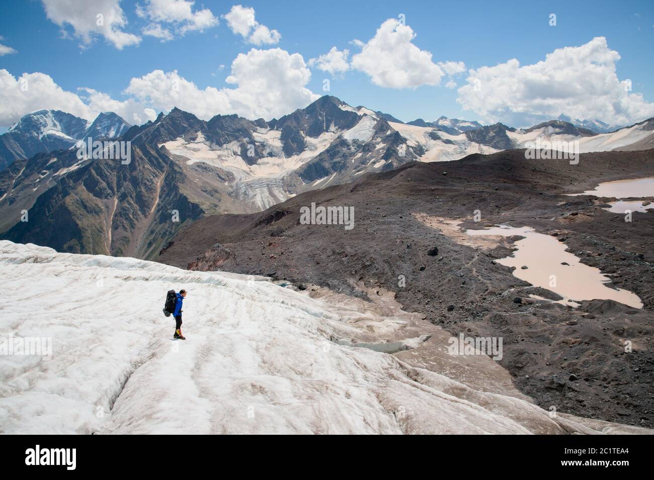 Un alpiniste avec un sac à dos marche dans des crampons le long d'un glacier poussiéreux avec des trottoirs dans les mains entre les fissures dans le Banque D'Images