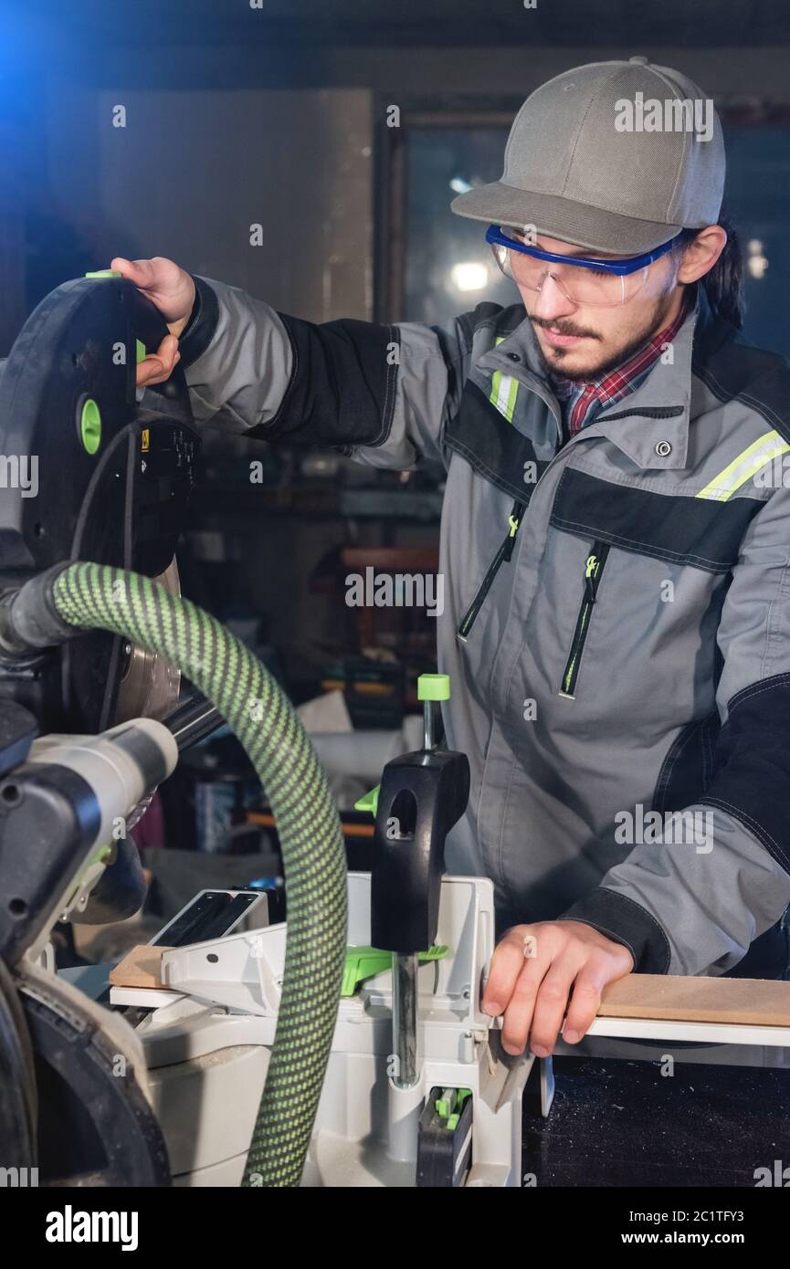 Jeune homme brunette portant une casquette dans une veste grise de  profession un charpentier coupe des planches de bois avec une scie  circulaire sur un travail Photo Stock - Alamy