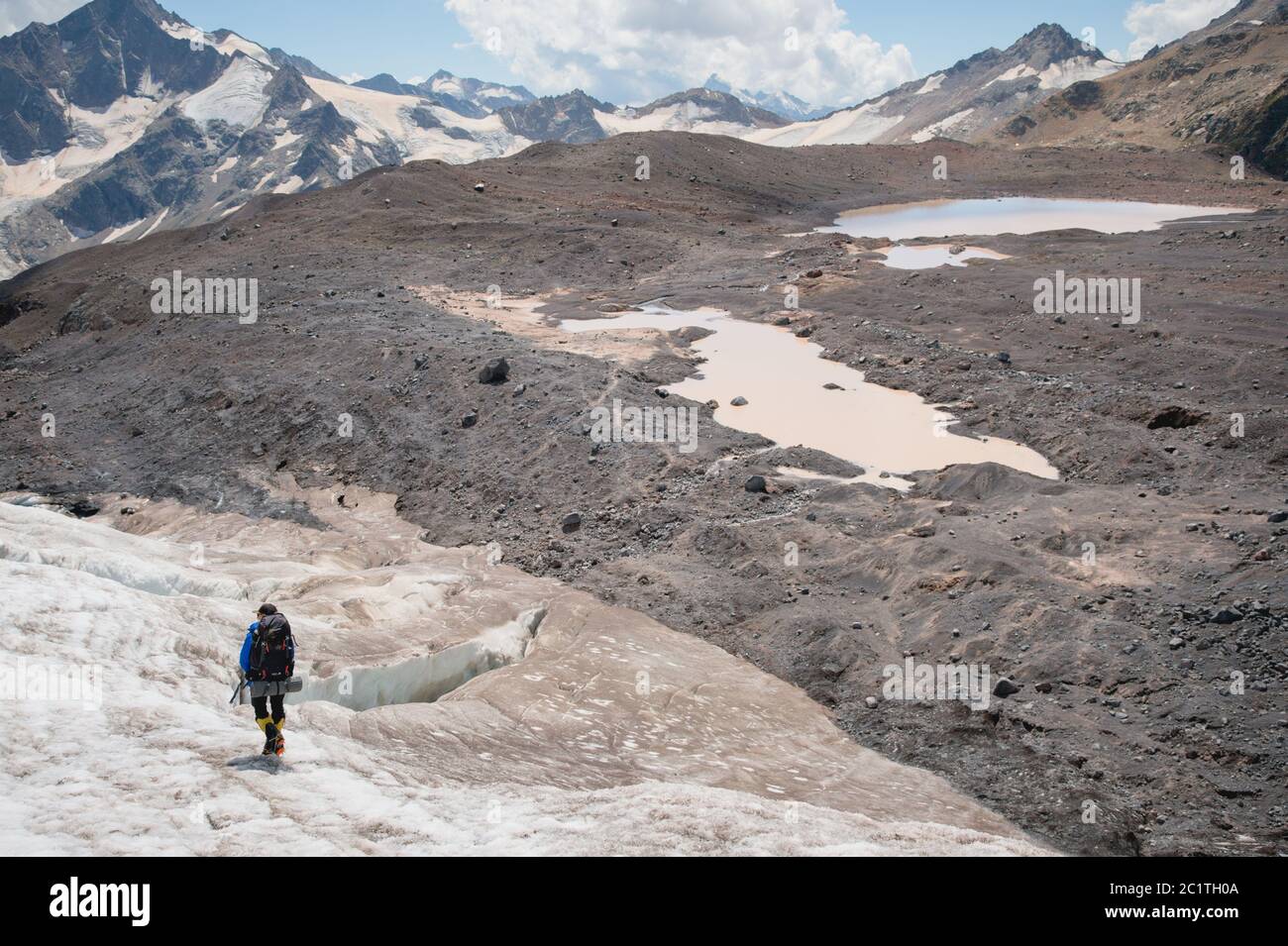 Un alpiniste avec un sac à dos marche dans des crampons le long d'un glacier poussiéreux avec des trottoirs dans les mains entre les fissures dans le Banque D'Images