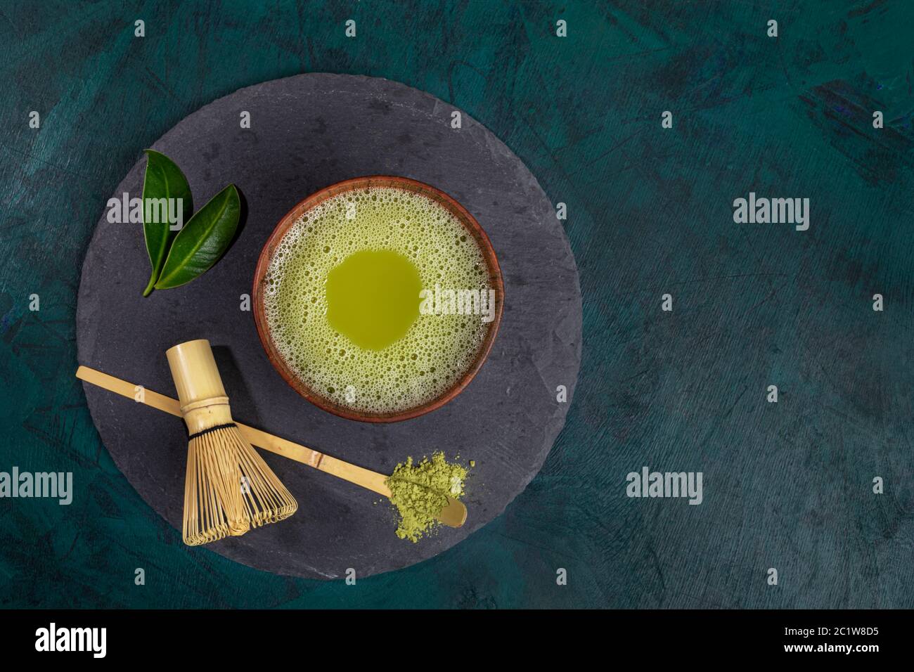 Tasse en bois avec thé vert matcha, fouet, cuillère et feuilles sur une table de service ronde de schiste sur fond émeraude. Le thé Matcha est un produit asiatique biologique sain Banque D'Images
