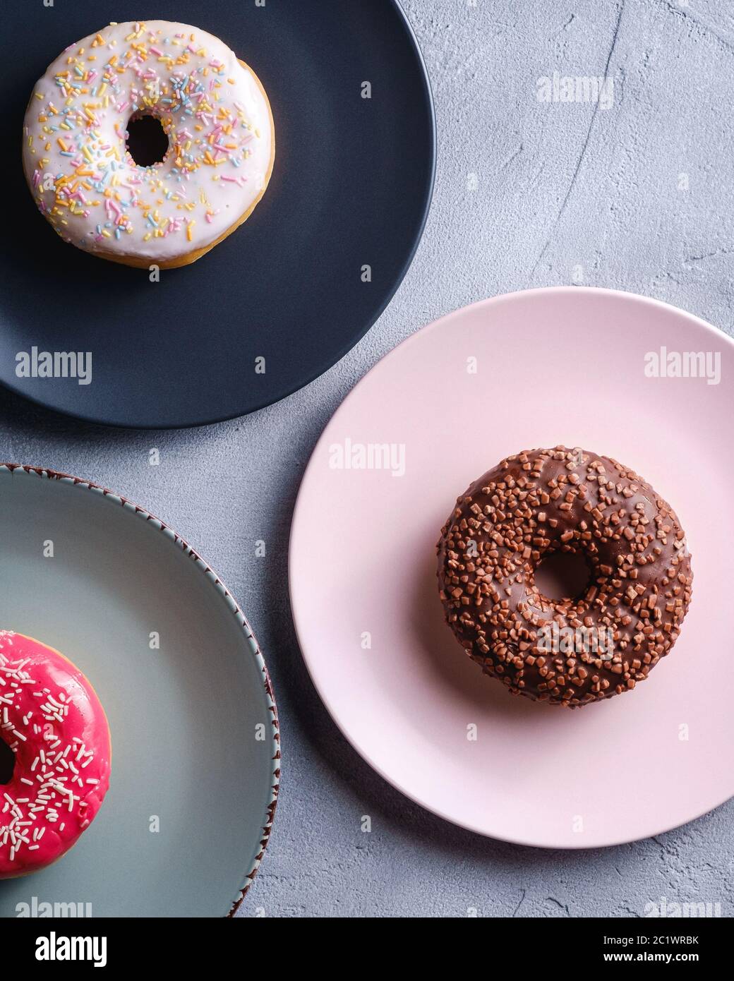 Trois beignets sur des assiettes, chocolat, beignets roses et vanille avec saupoudrés, dessert glacé sucré sur fond texturé en béton, vue du dessus Banque D'Images