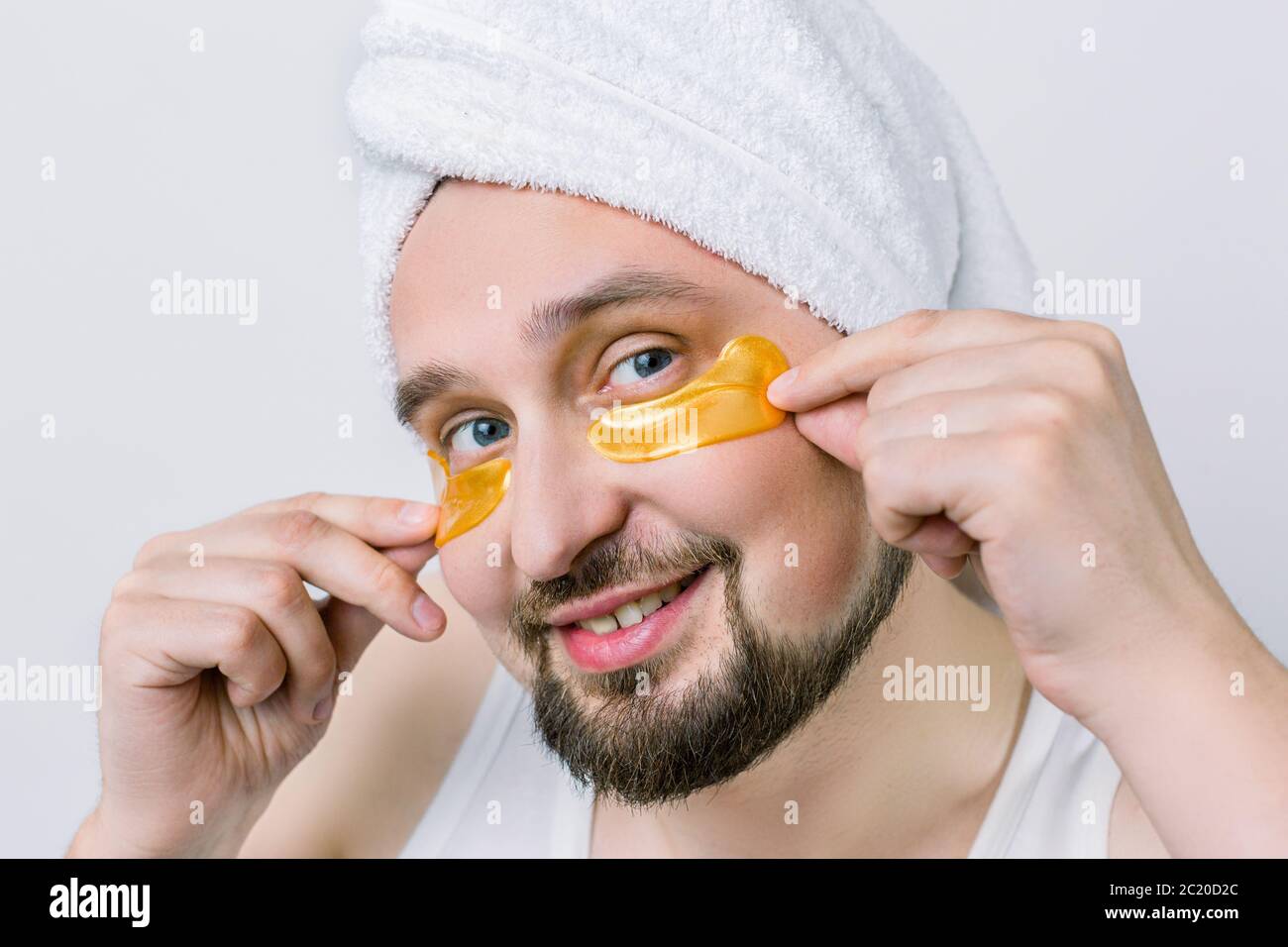 Homme barbu souriant avec une serviette blanche sur la tête et des taches dorées sous les yeux, prenant soin de sa peau de visage regardant l'appareil photo sur fond blanc Banque D'Images