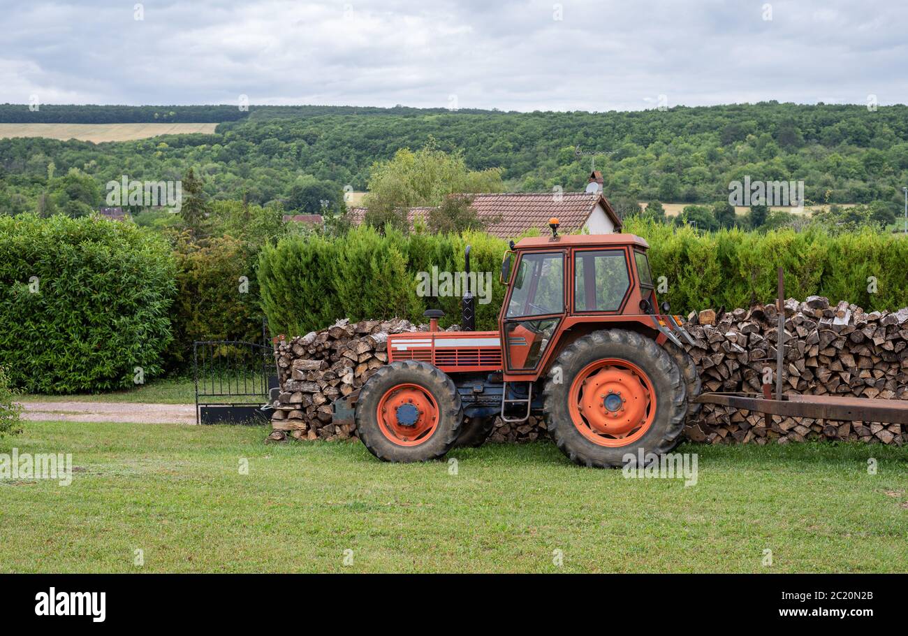 Tracteur rouge stationné dans un jardin devant une pile de bois coupé. Paysage de campagne. Banque D'Images