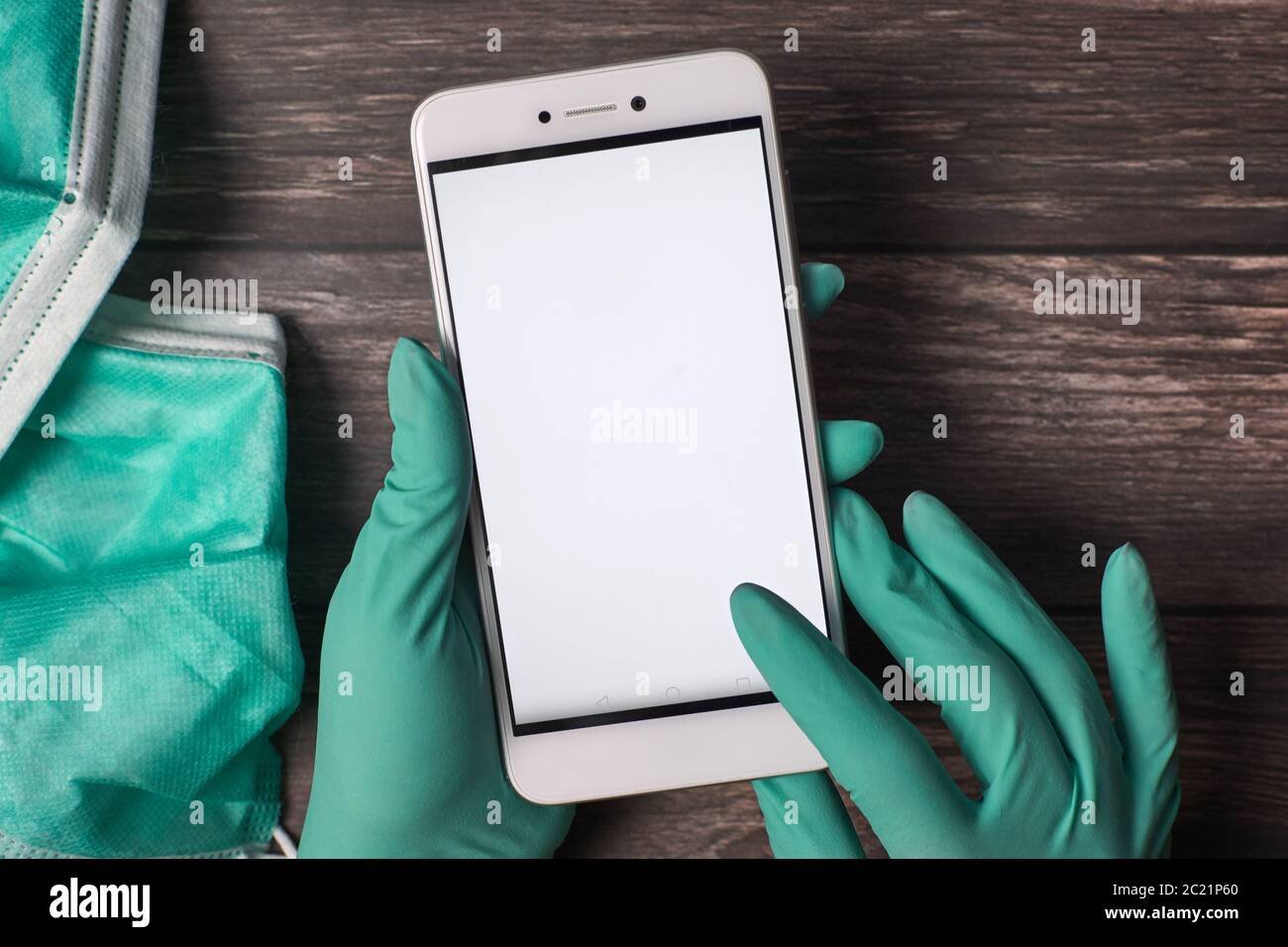 Mains du médecin portant des gants chirurgicaux et utilisant un smartphone avec écran blanc blanc blanc, masques sur une table en bois. Banque D'Images