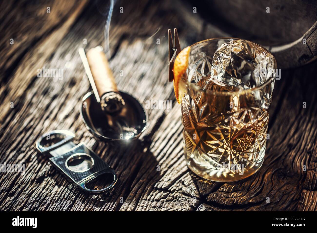 Verre ornemental plein de whisky et de glace sur un bois vintage à côté d'un coupe-cigare et d'un cigare en feu Banque D'Images