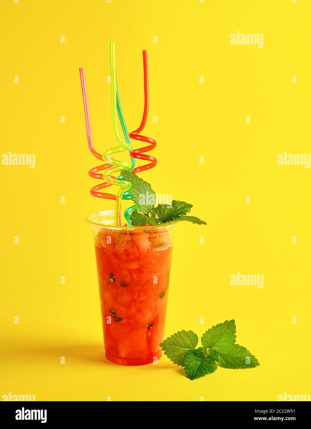 De smoothie pastèque rouge mûr dans une tasse en plastique, lot de tubes colorés et une feuille de menthe sur fond jaune Banque D'Images