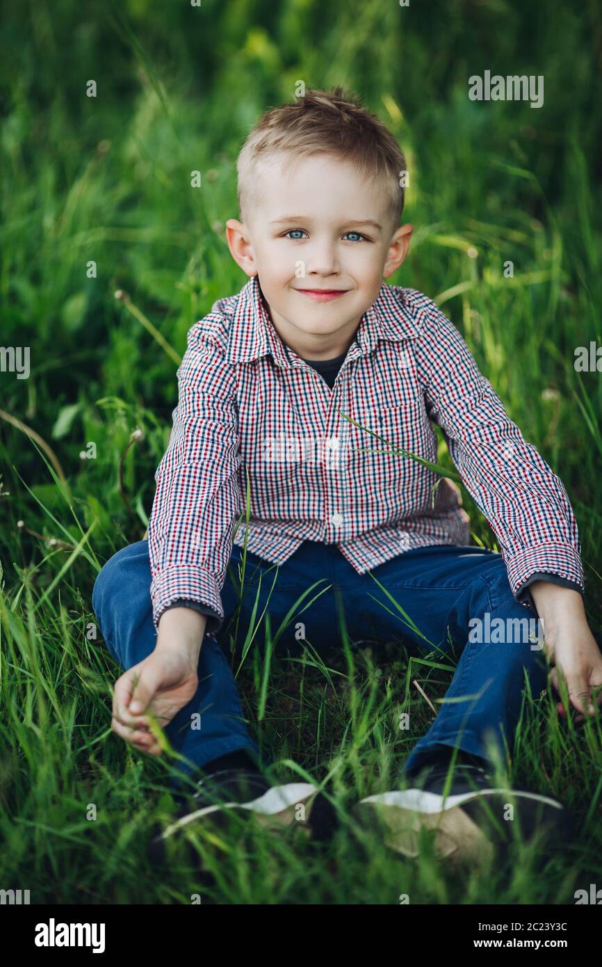 Portrait de petit garçon blond élégant assis dans un parc parmi l'herbe. Banque D'Images