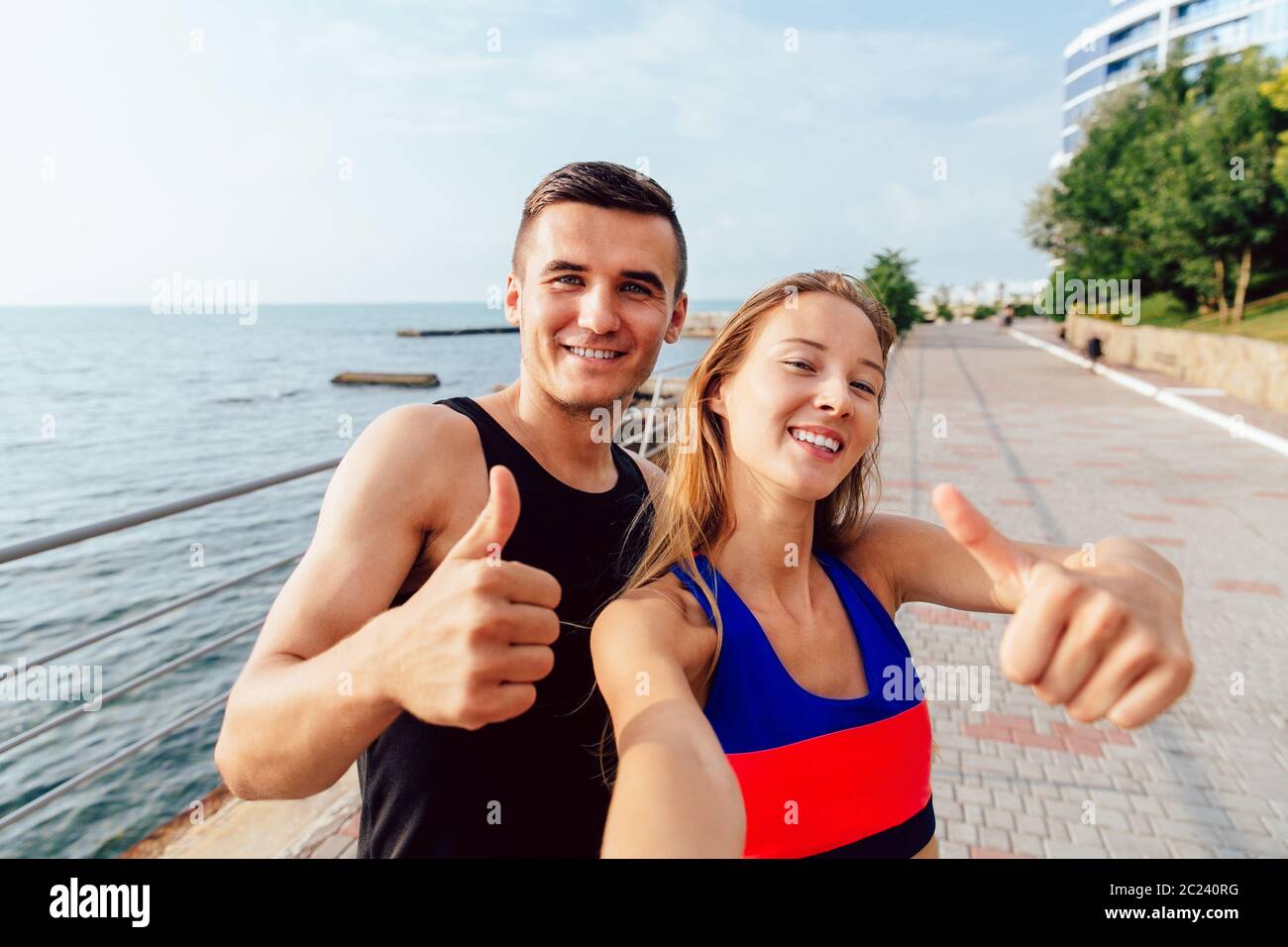 Sourire de la sportive et sportif de sportswear montrant un pouce vers le haut tout en tenant un lors de l'entraînement selfies sur le quai, près de la mer. Banque D'Images