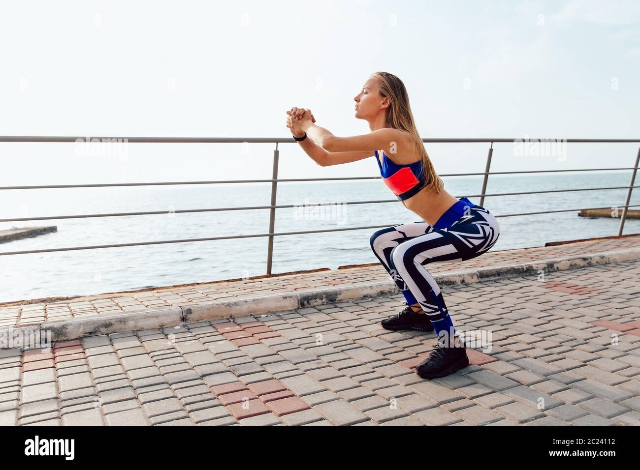 Vue latérale d'assez grande sportive faisant des squats, des exercices sportifs pour le corps au cours de la formation sur le quai, près de la mer. Banque D'Images