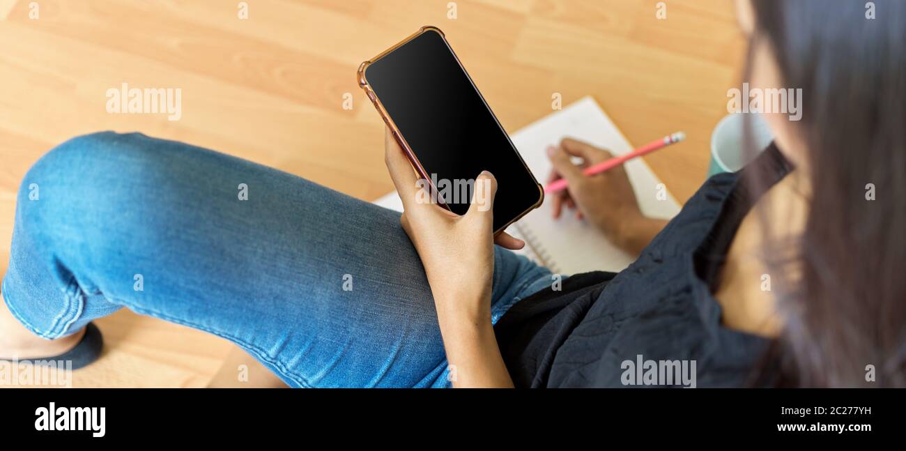 Photo rognée d'une femme Prenez note sur un ordinateur portable vierge tout en regardant des informations sur un smartphone Banque D'Images