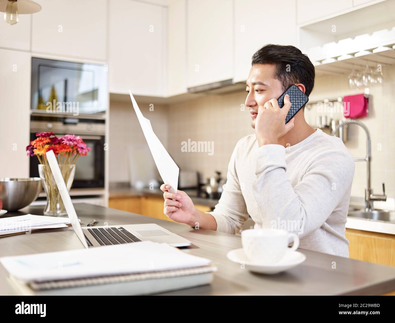 jeune homme d'affaires asiatique assis cuisine comptoir travaillant à la maison regardant un reportage parlant sur téléphone mobile Banque D'Images