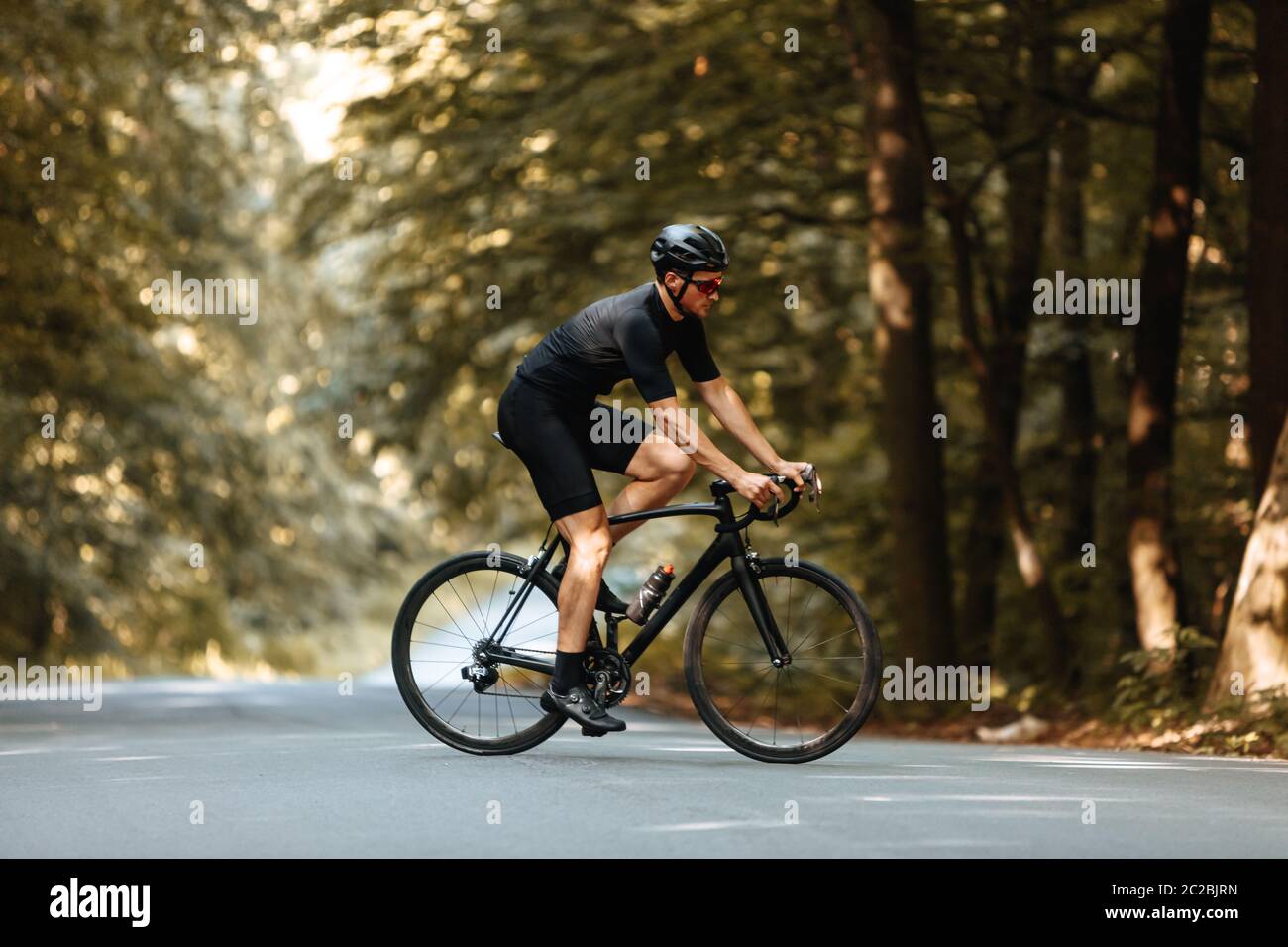 Vue latérale de l'homme actif dans des vêtements de sport à vélo noir avec de beaux arbres verts autour. Cycliste barbu dans un casque de protection et des lunettes Banque D'Images