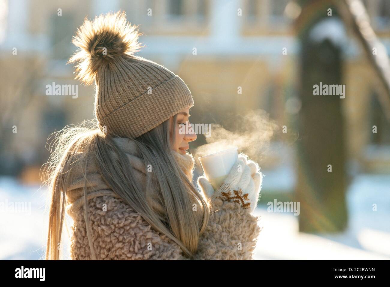 Mignon jeune femme caucasienne en fourrure beige, chapeau avec pompon, écharpe et moufles blanches tenant la vapeur tasse blanche de thé chaud ou de café, à l'extérieur au soleil Banque D'Images