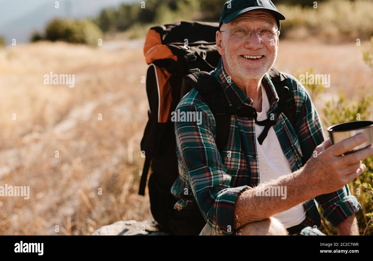 Un randonneur mâle senior ayant un café sur le sentier de randonnée. Un randonneur prend du repos et boit du café frais, regarde l'appareil photo et sourit. Banque D'Images