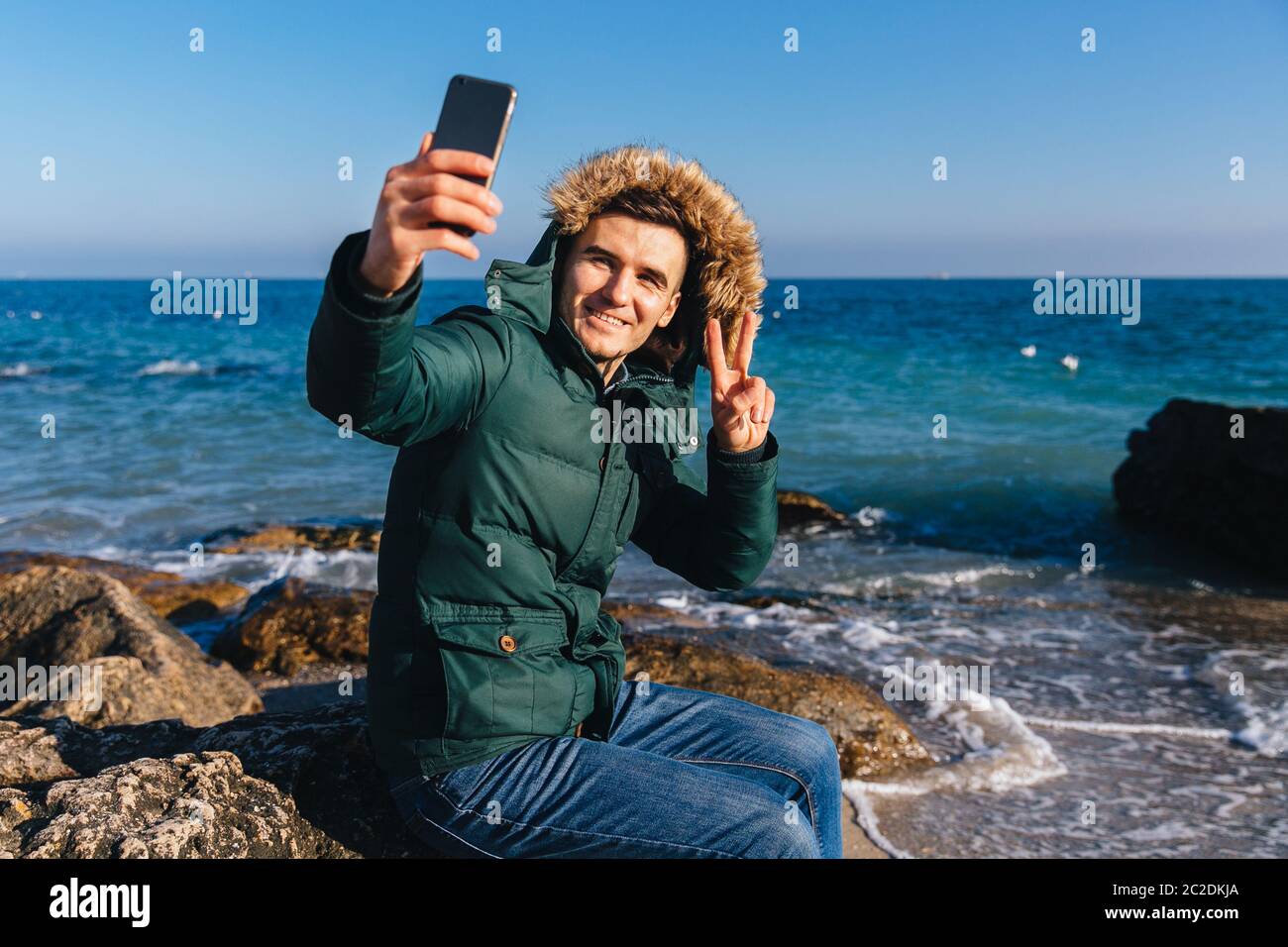 Heureux bel homme prend un smartphone sur selfies, montrant geste de paix sur le fond de la mer. Vêtus de veste chaude avec capuchon de fourrure. Banque D'Images