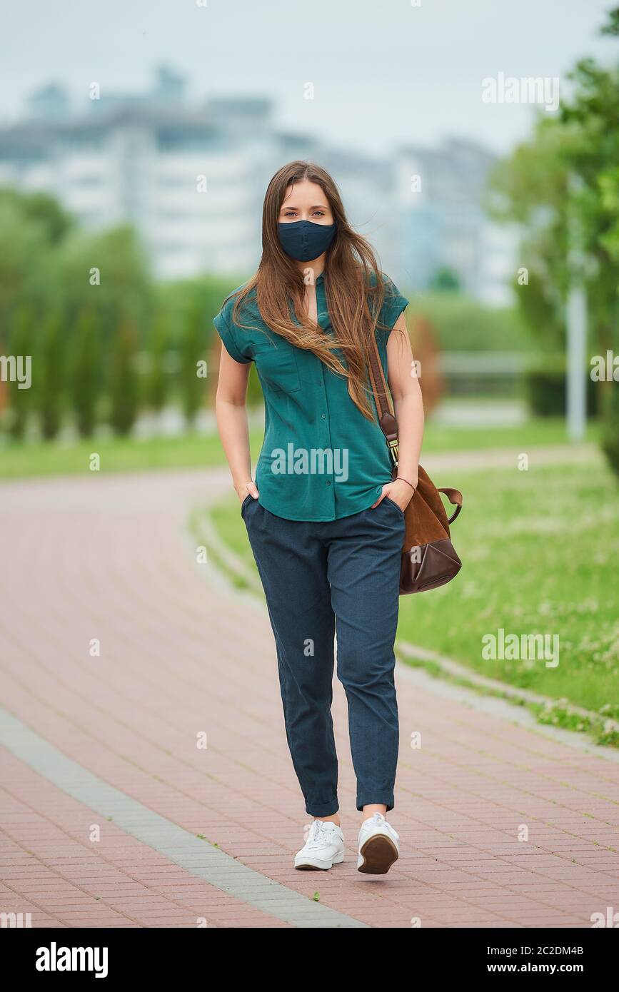 Une jeune femme dans un masque médical marche les mains en se portant dans les poches de pantalons dans le parc. Fille gardant la distance sociale Banque D'Images