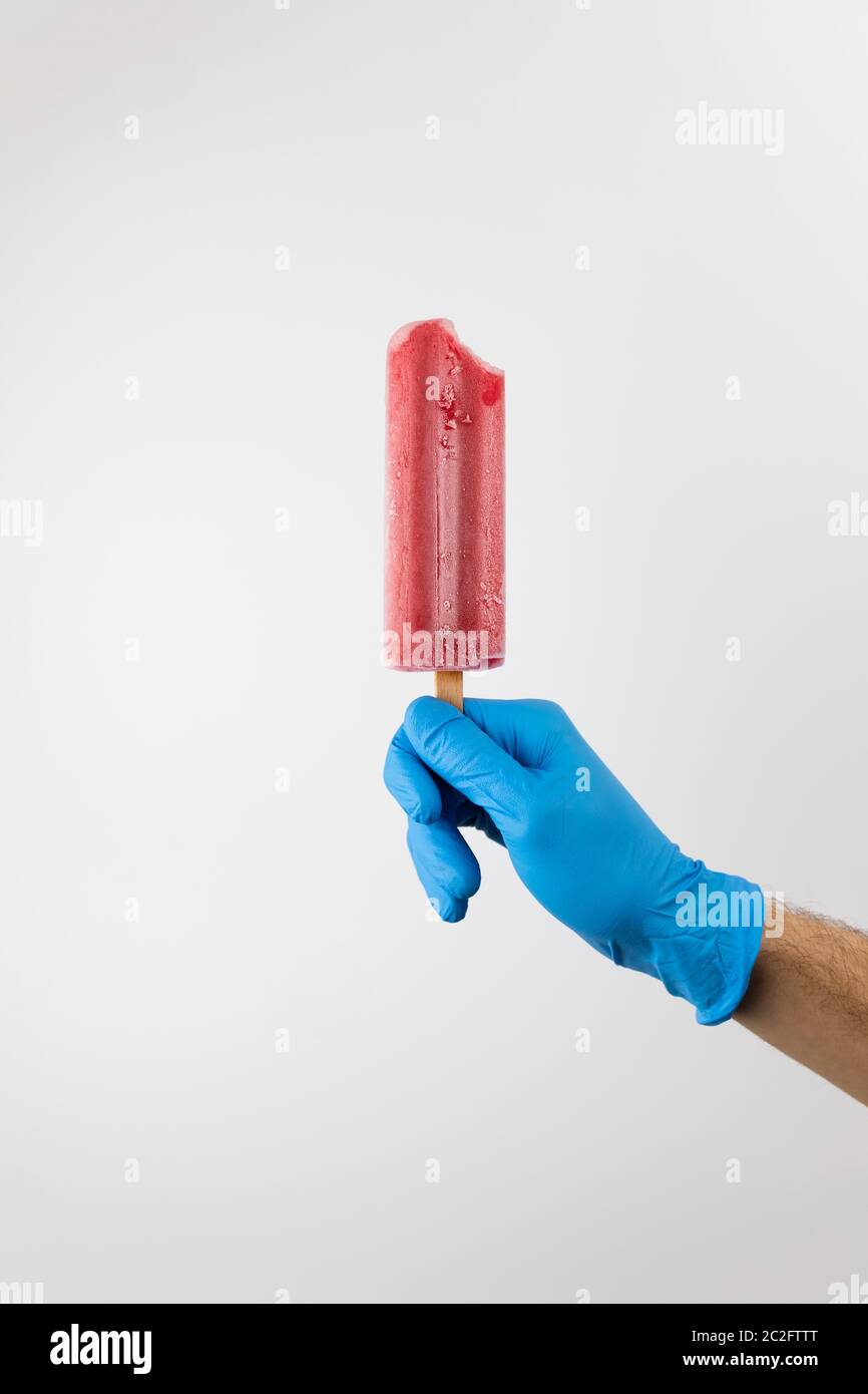 Main de l'homme tenant la glace rouge de la fraise popsicle, portant des gants bleus sur fond blanc. Prévention de la pandémie du coronavirus Banque D'Images