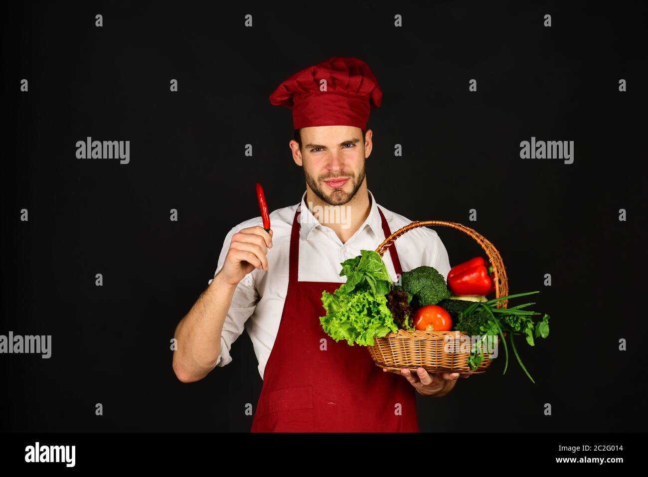 Le chef en uniforme bordeaux tient le piment rouge à la main. Concept de  repas naturel et épicé. Homme avec barbe et légumes sur fond noir. Cuisinez  avec un visage souriant, du