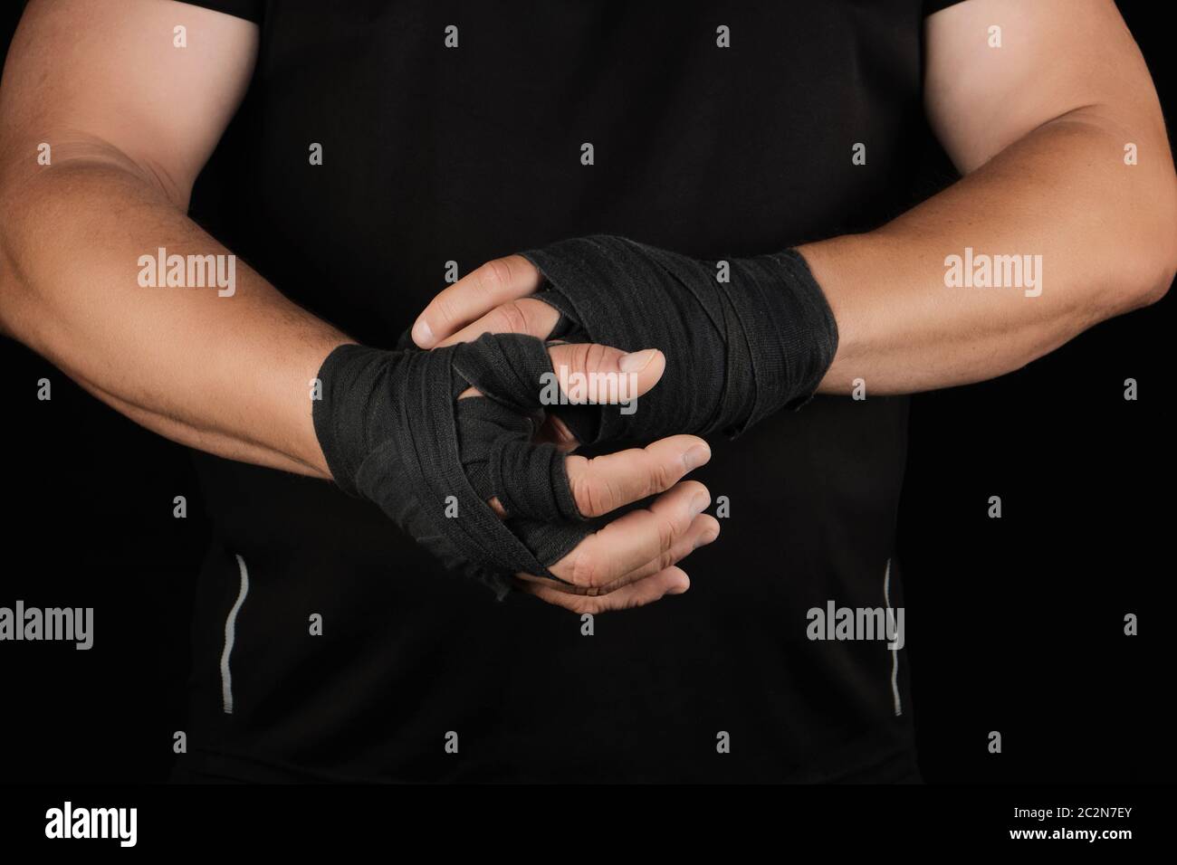 les athlètes adultes se tiennent dans des vêtements noirs et enveloppent ses mains dans un bandage élastique en textile noir avant l'entraînement, le fond noir, le corps musculaire Banque D'Images