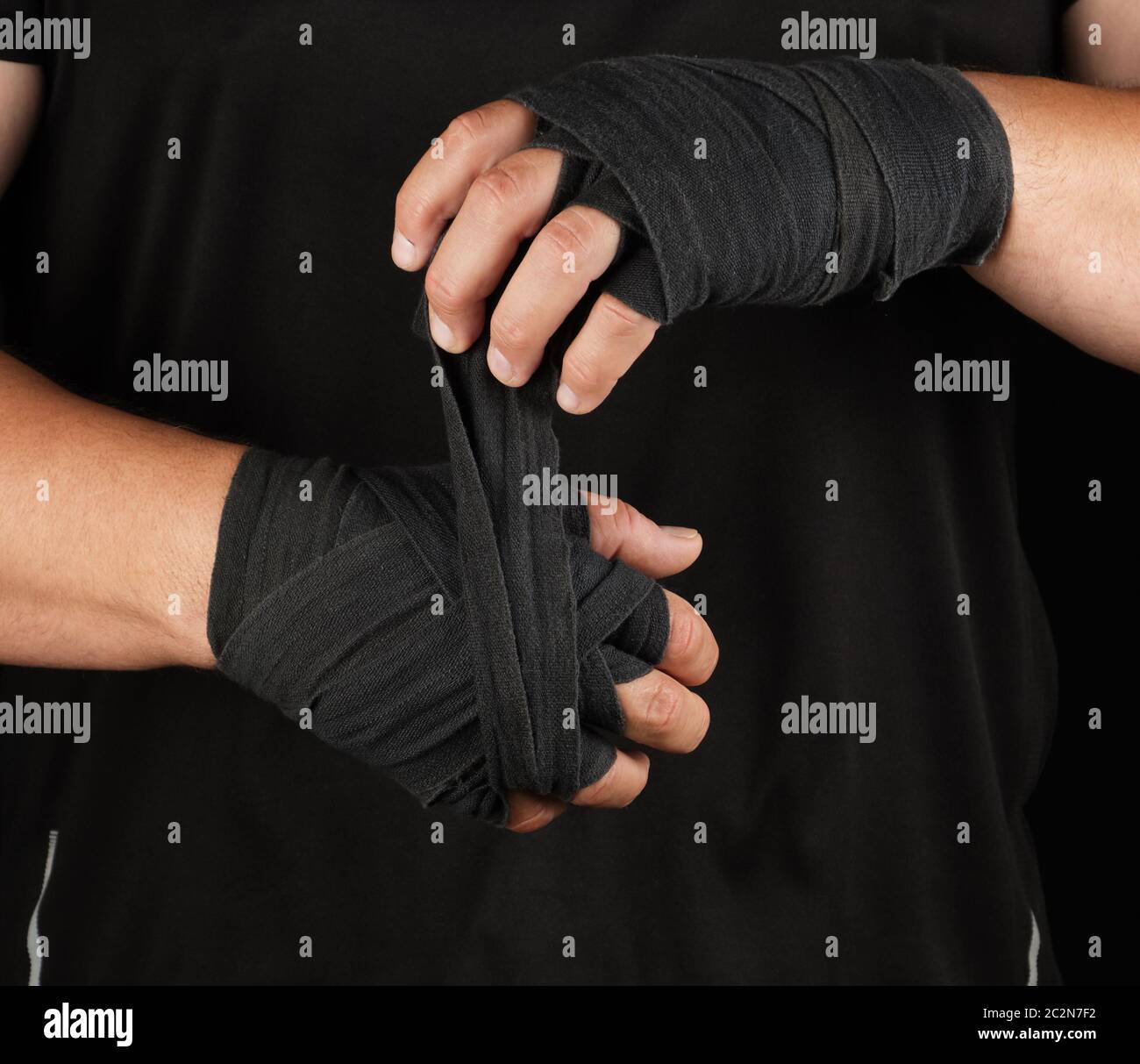 les athlètes adultes se tiennent dans des vêtements noirs et enveloppent ses mains dans un bandage élastique en textile noir avant l'entraînement, le fond noir, le corps musculaire Banque D'Images